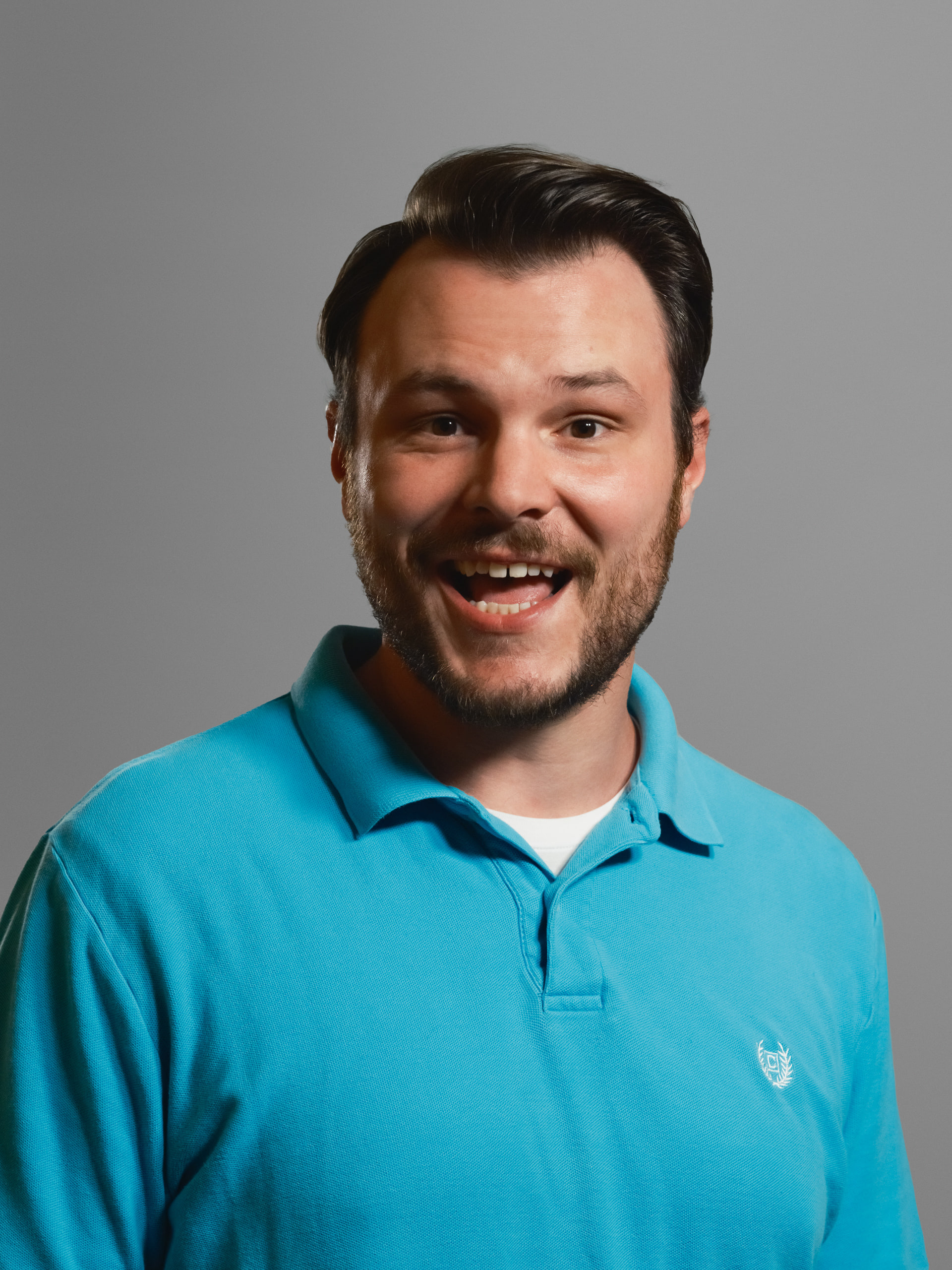 A studio portrait from a team portrait day: a man in a teal polo against a plain gray ground
