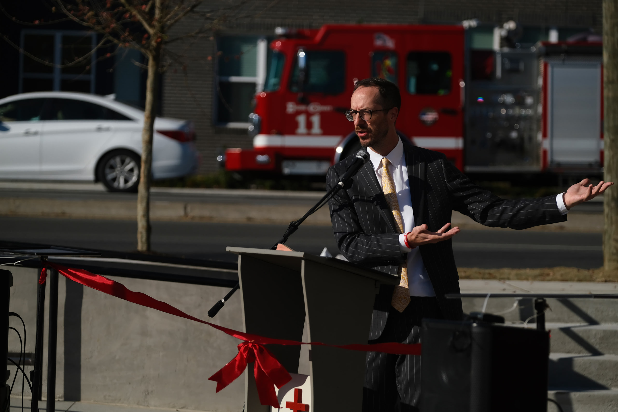 A speaker with his arms out at a podium beside a red ribbon, a fire truck parked behind him, at a building dedication