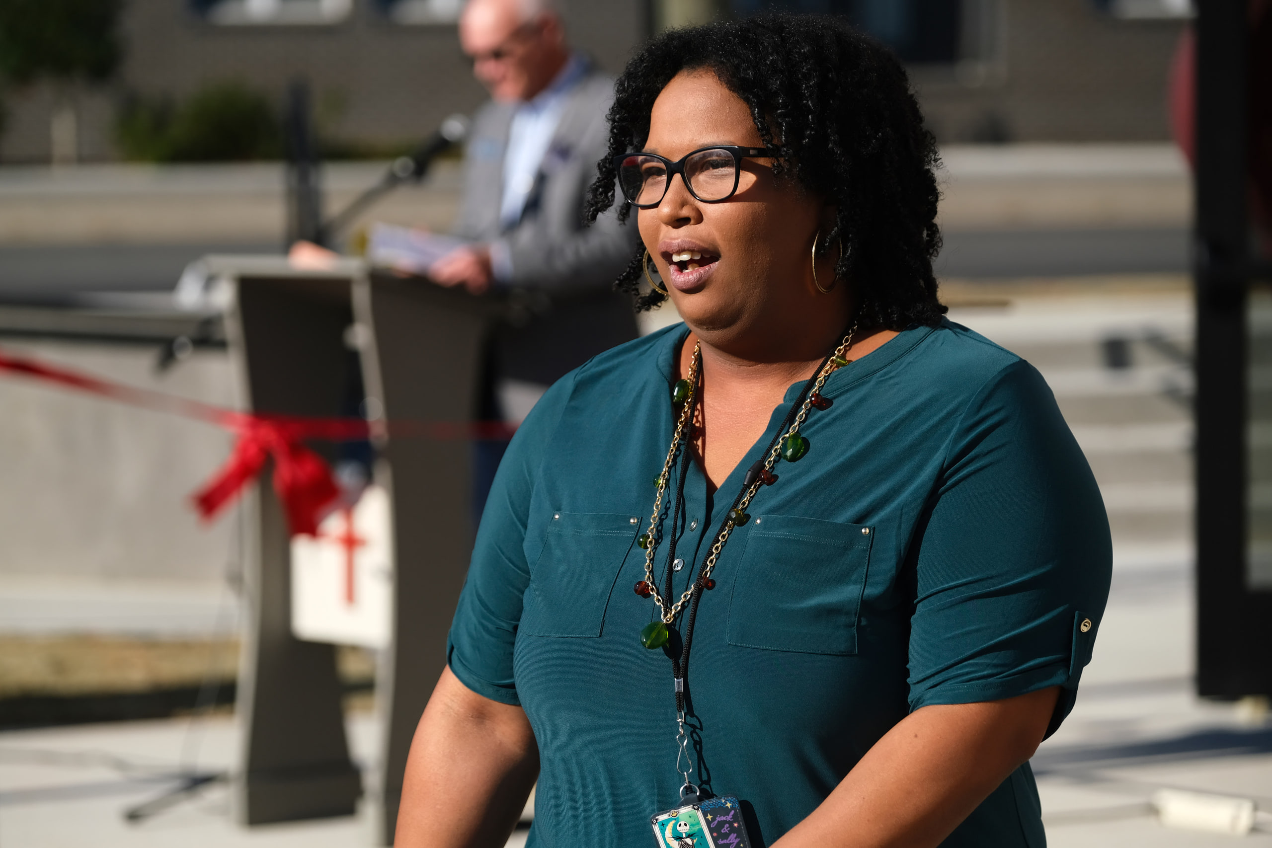 Woman speaking, teal, the crowd behind at a nonprofit building dedication.