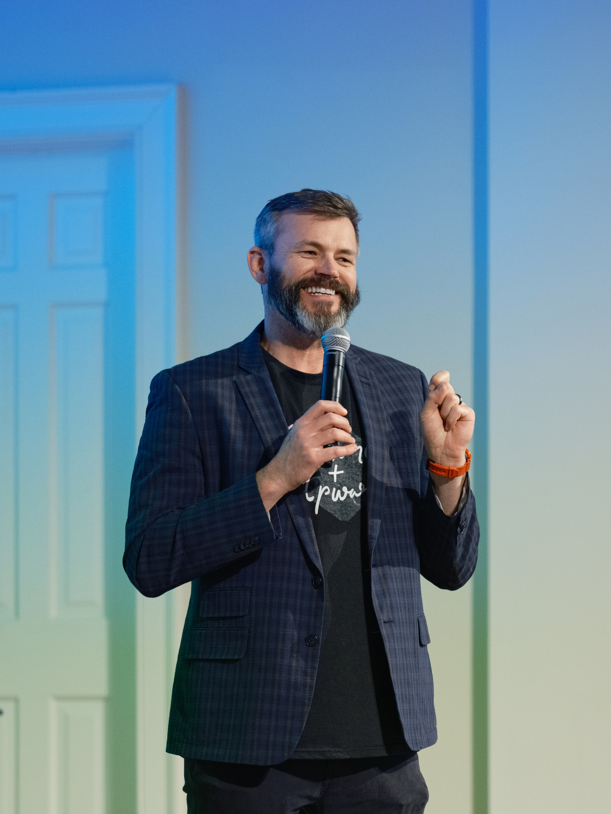 A speaker in a blazer holding a microphone on stage, smiling at the audience