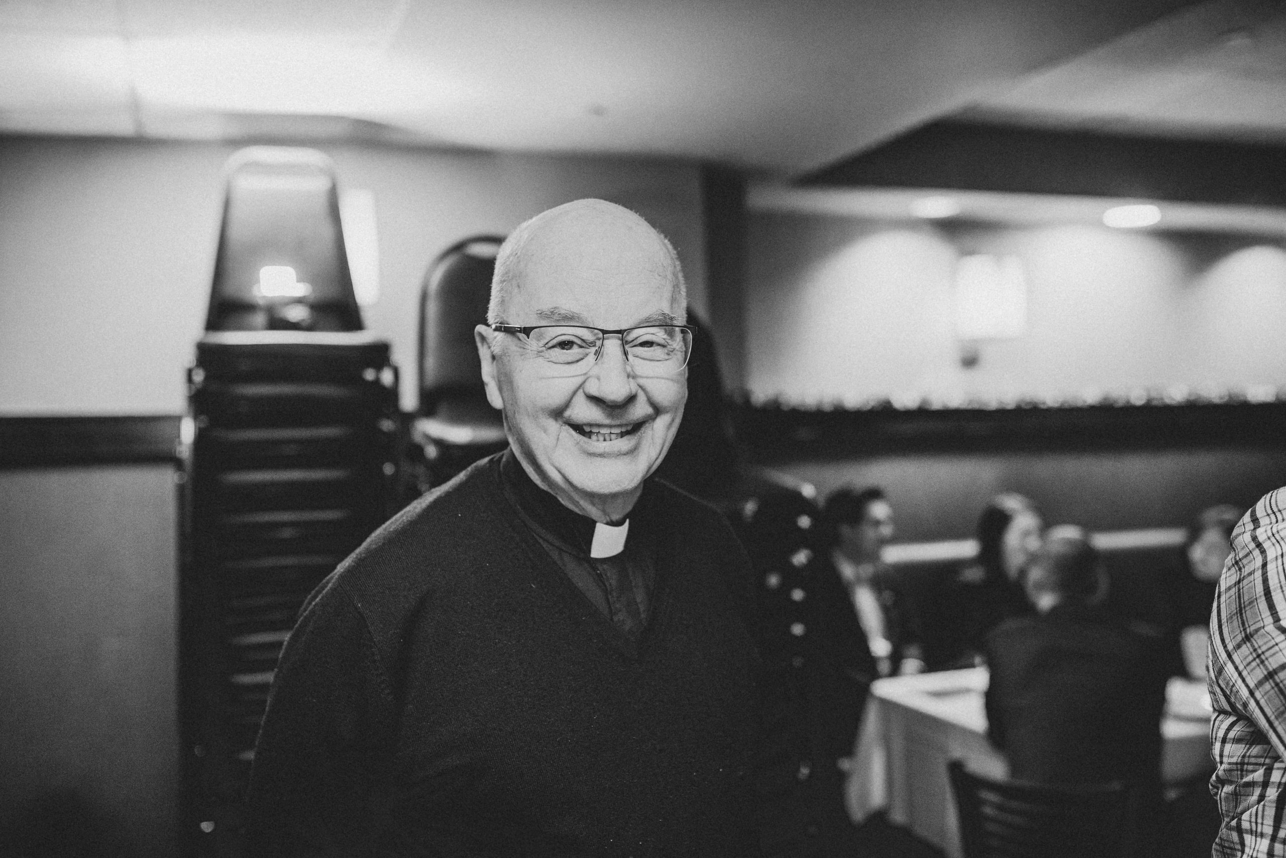 Black and white photo of a smiling older man with glasses and a clerical collar in a banquet room, with stacked chairs and seated guests blurred behind him.