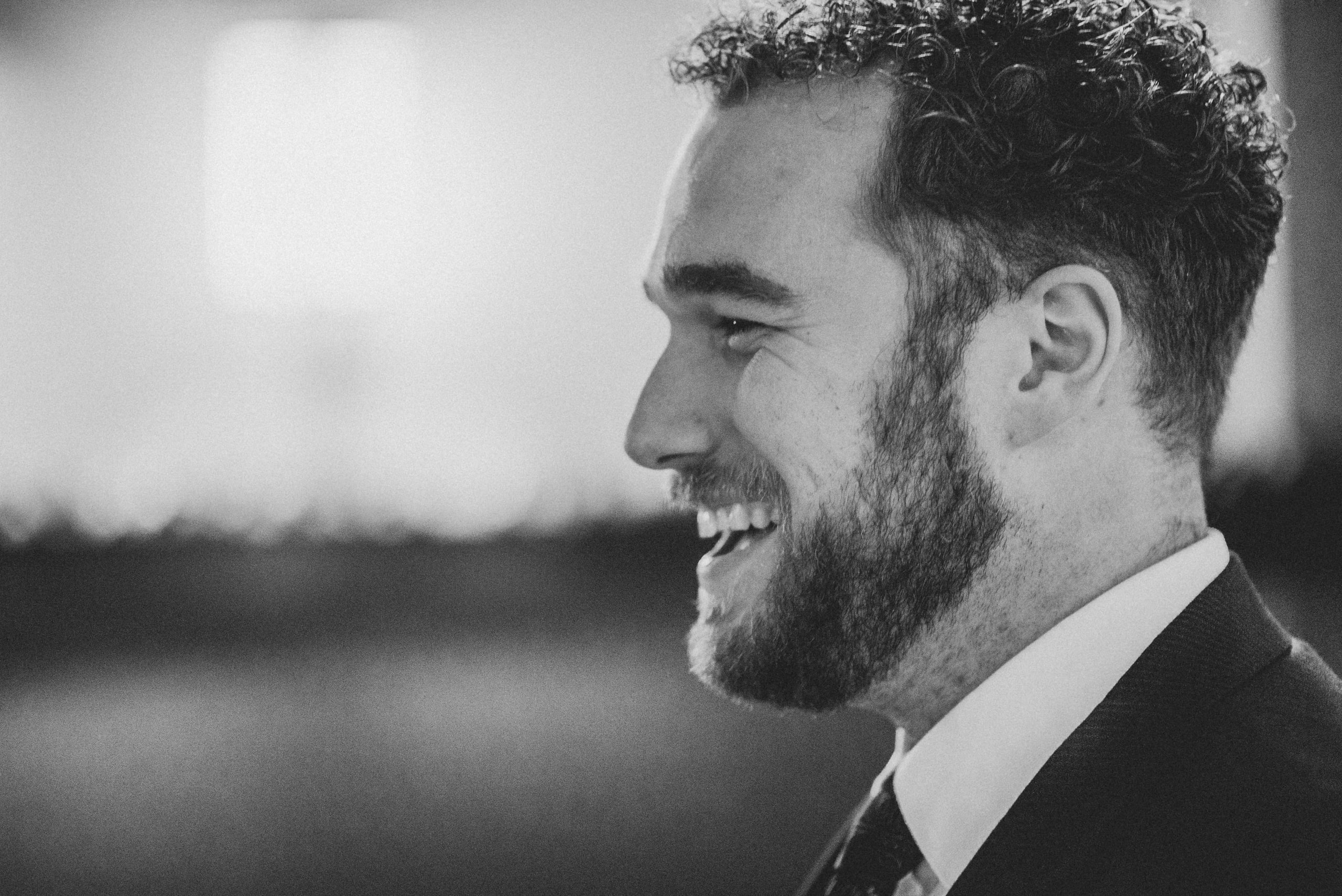 Black and white profile of a bearded man with curly hair in a dark suit and tie laughing, the room behind him blurred to soft light.