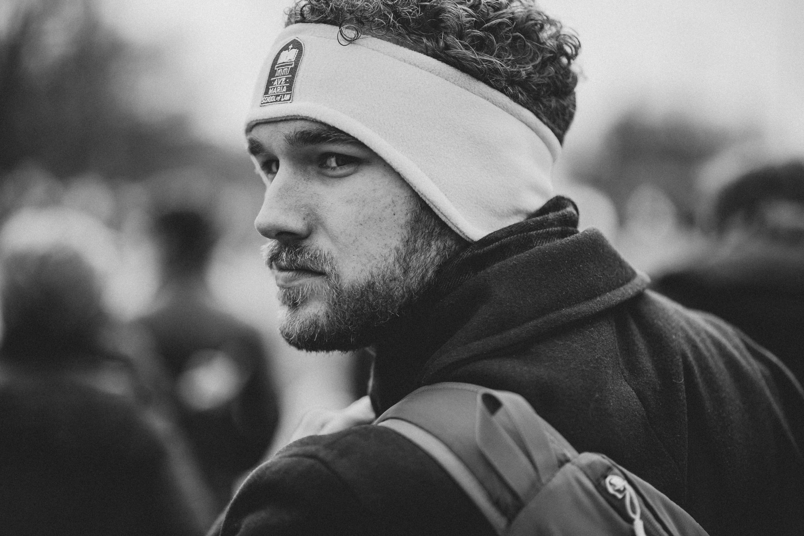 Black and white close portrait of a bearded young man with curly hair and a light fleece headband glancing back over his shoulder at the camera, with a blurred crowd behind him.