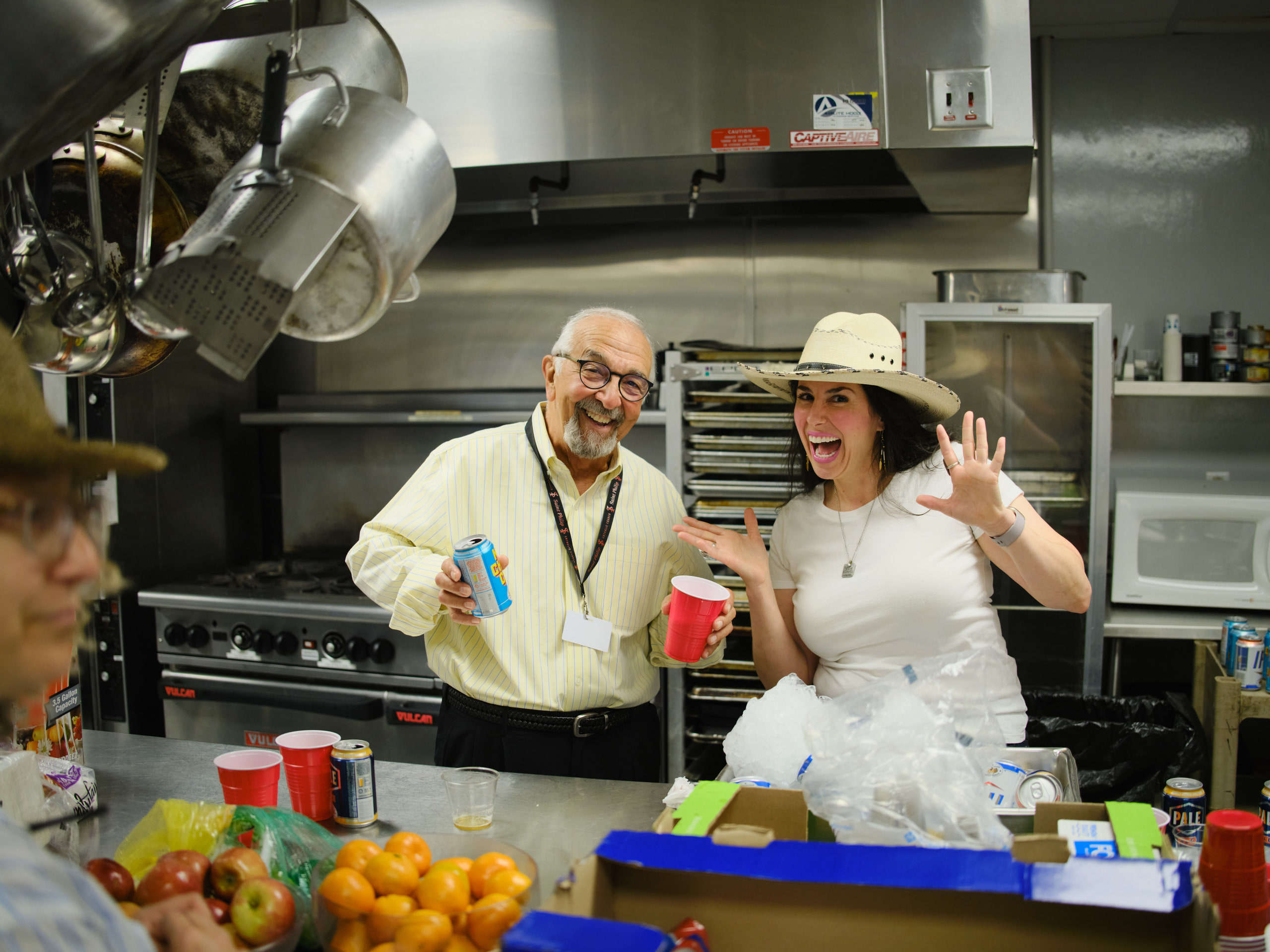 Two in the kitchen with hats, laughing at a community event.