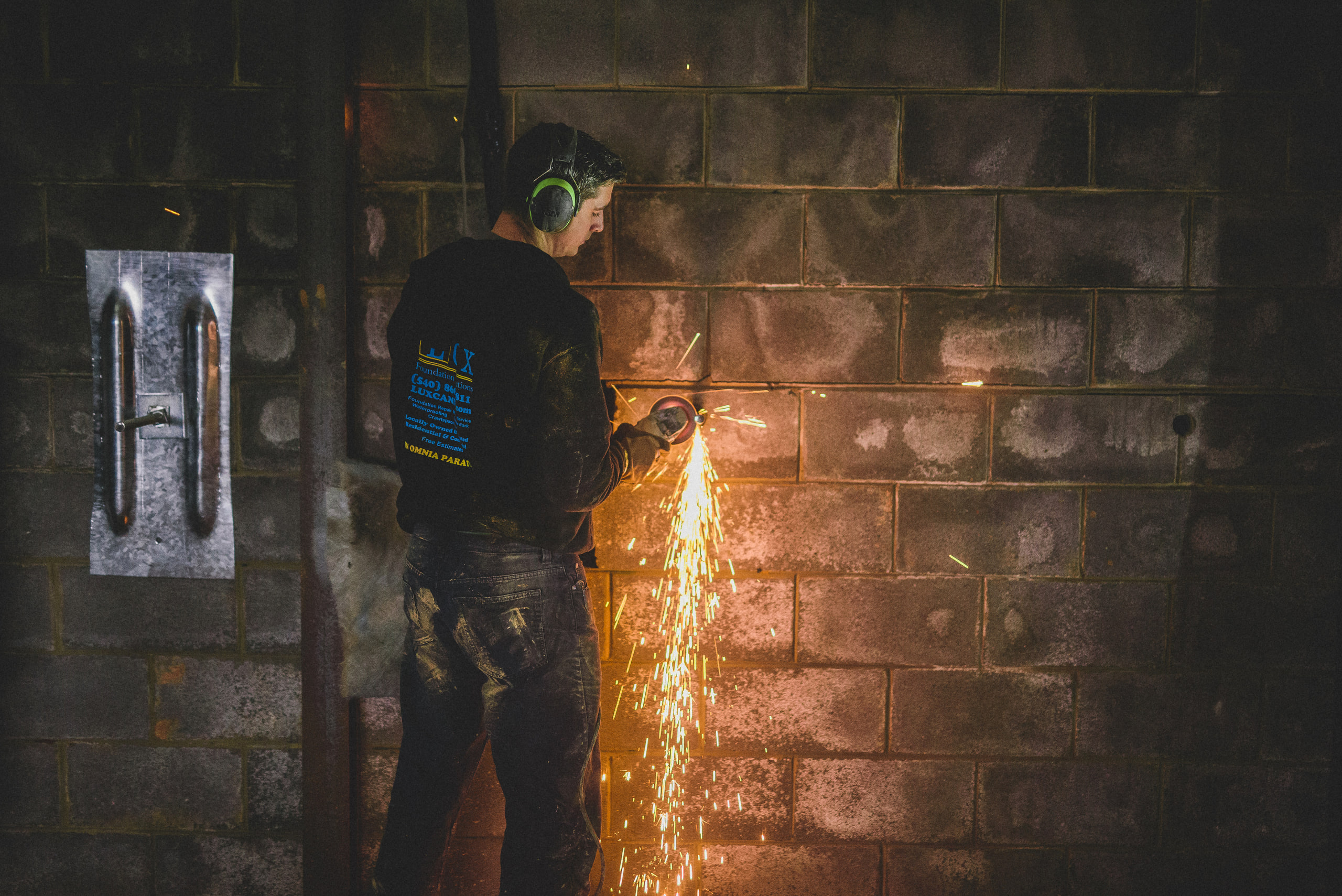 A worker in ear protection cutting into a block wall with a grinder, sparks spraying, on a foundation repair job