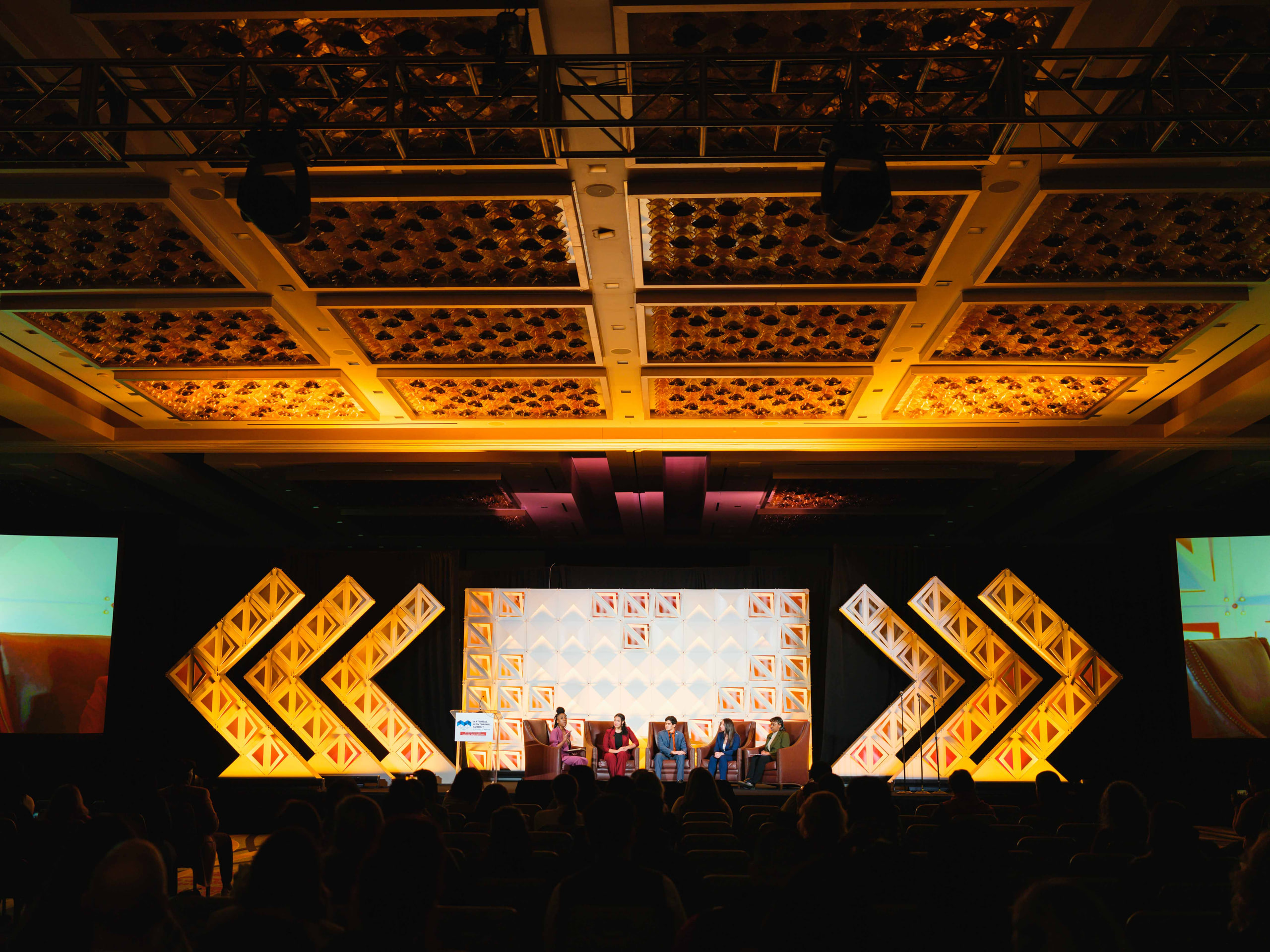 A wide view of a ballroom under a gold-lit ceiling, a panel small on the stage between bright chevron screens.
