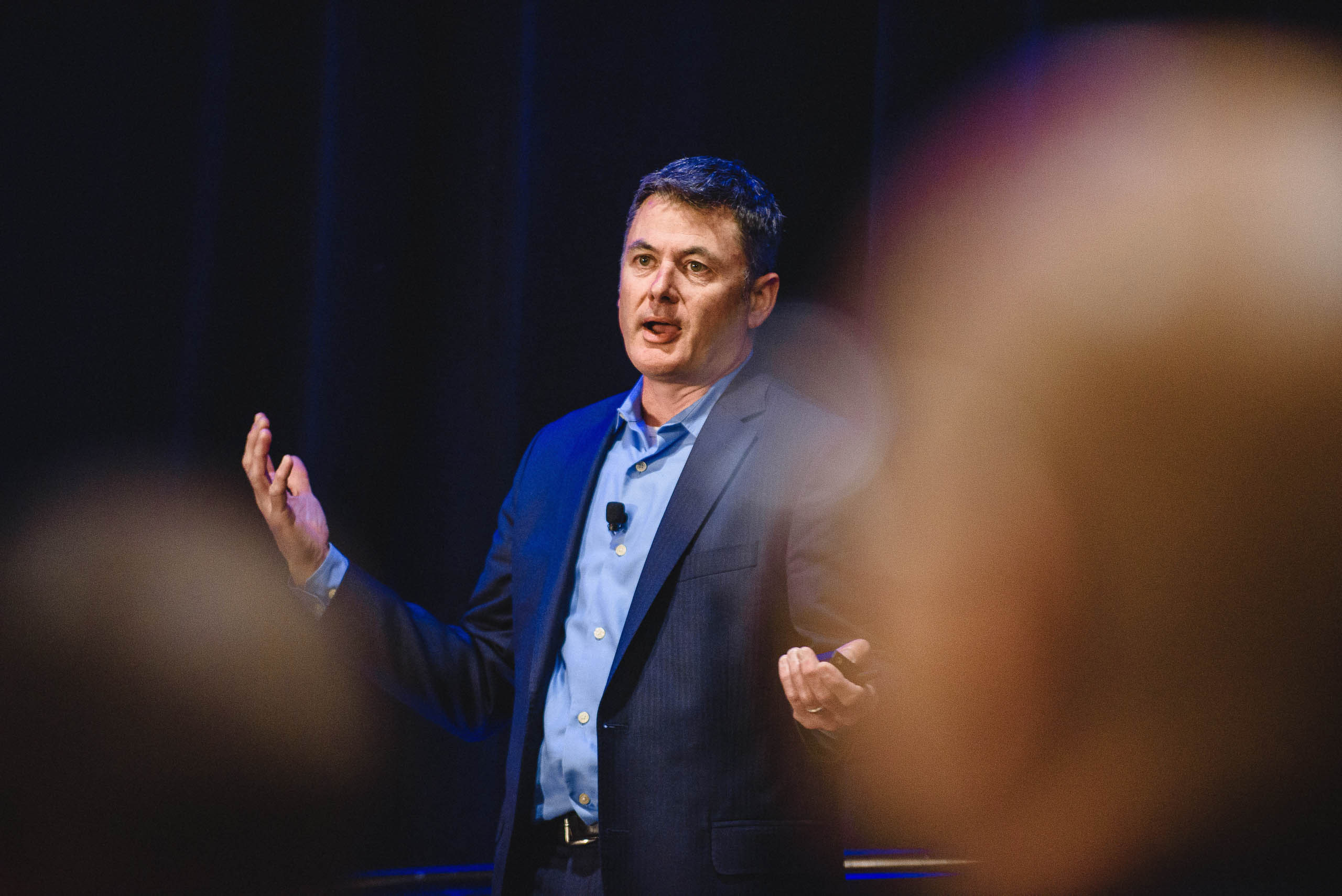 A keynote speaker in a navy blazer gesturing on a dark blue stage, framed between blurred audience members.