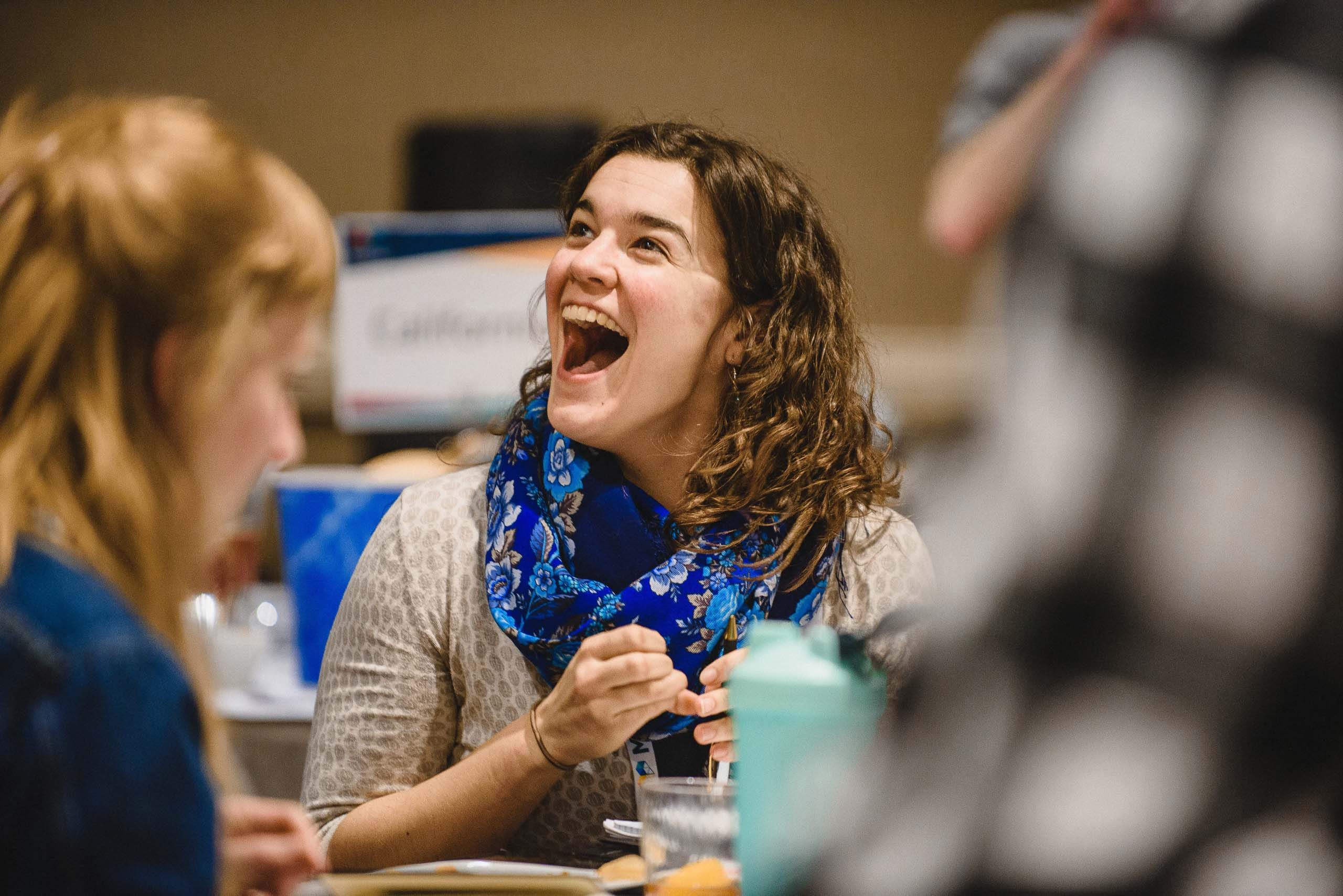 A conference attendee in a blue patterned scarf laughing open-mouthed at her table during a national summit session.