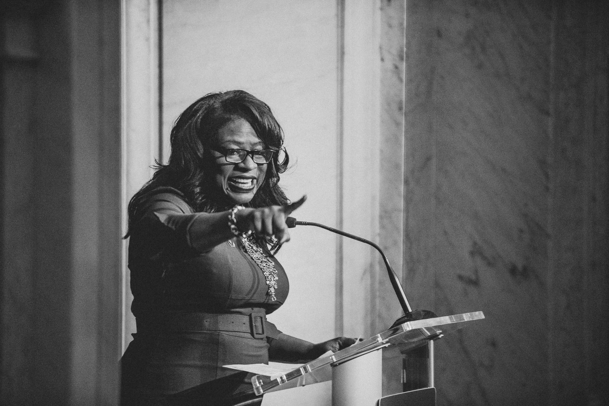 Black and white: a woman at the lectern pointing straight at the camera, laughing, a microphone beside her.