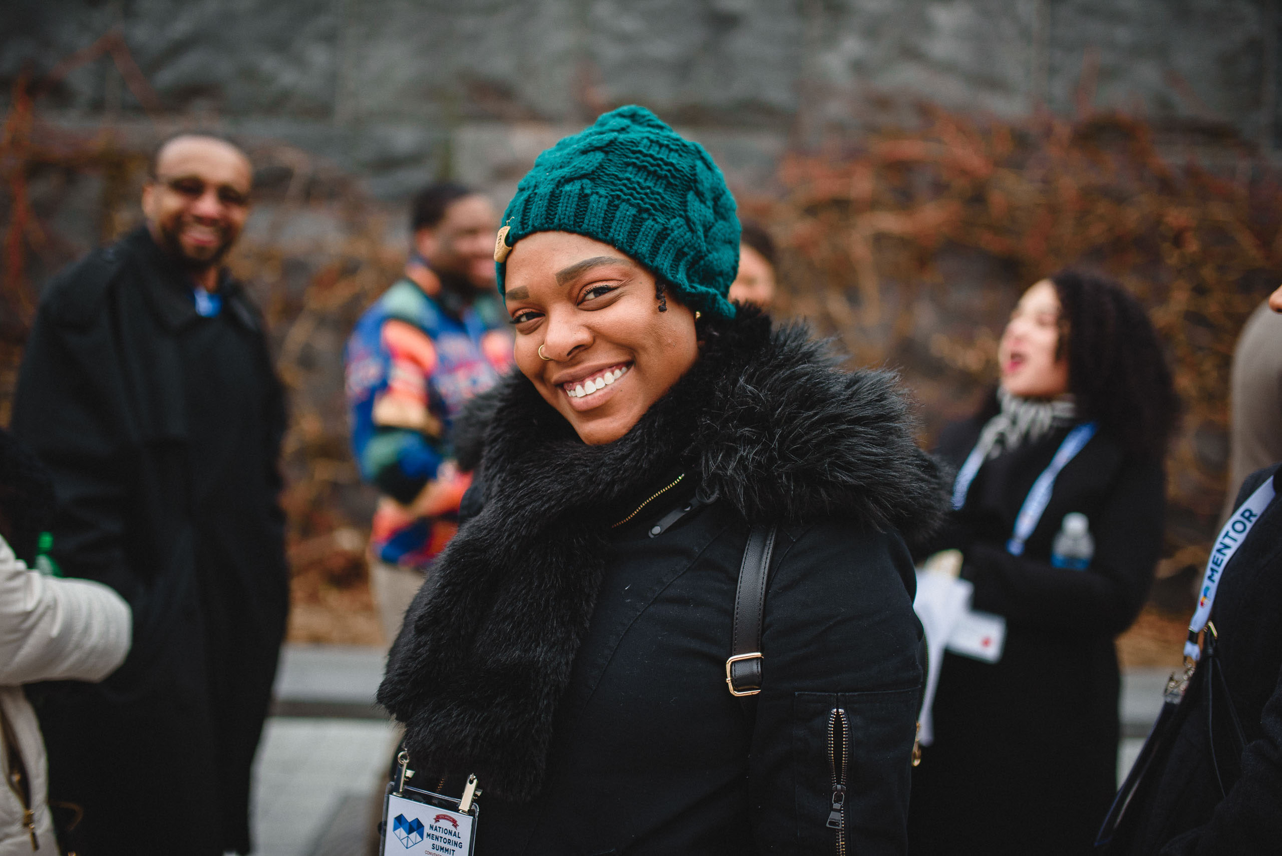 A woman in a green knit hat and black winter coat laughing at the camera, with other attendees behind her.