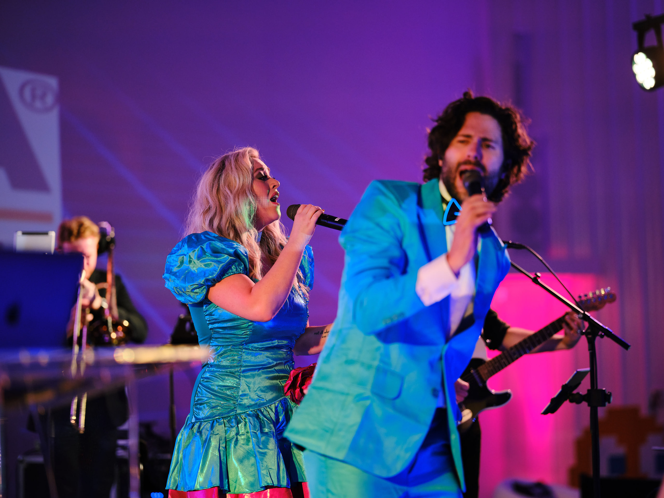 A singer in a blue party dress and a singer in a bright blue suit performing together on a lit stage during an evening gala