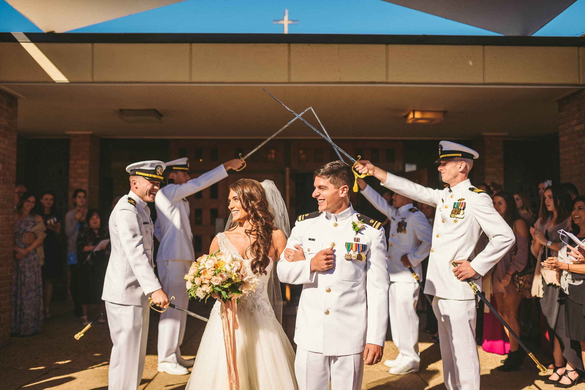 A bride and groom in naval dress whites walk out of a church under an arch of raised swords, laughing, with guests watching