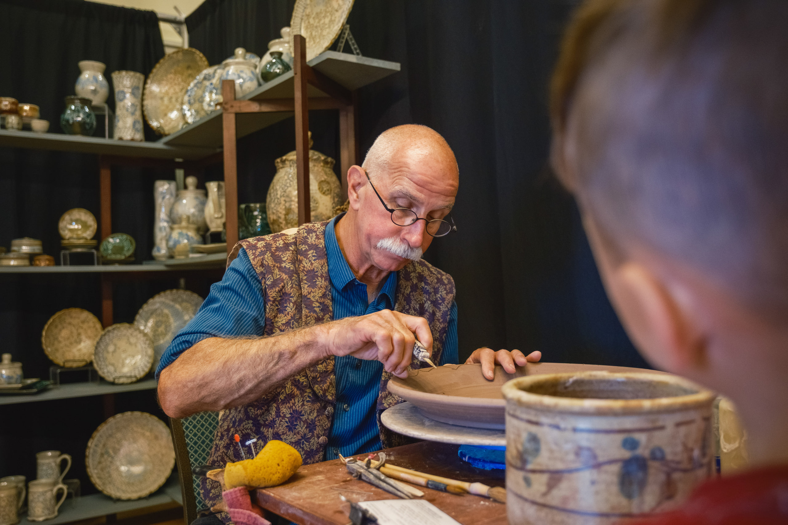 A potter in glasses and a patterned vest painting a plate at his wheel, shelves of stoneware behind him.