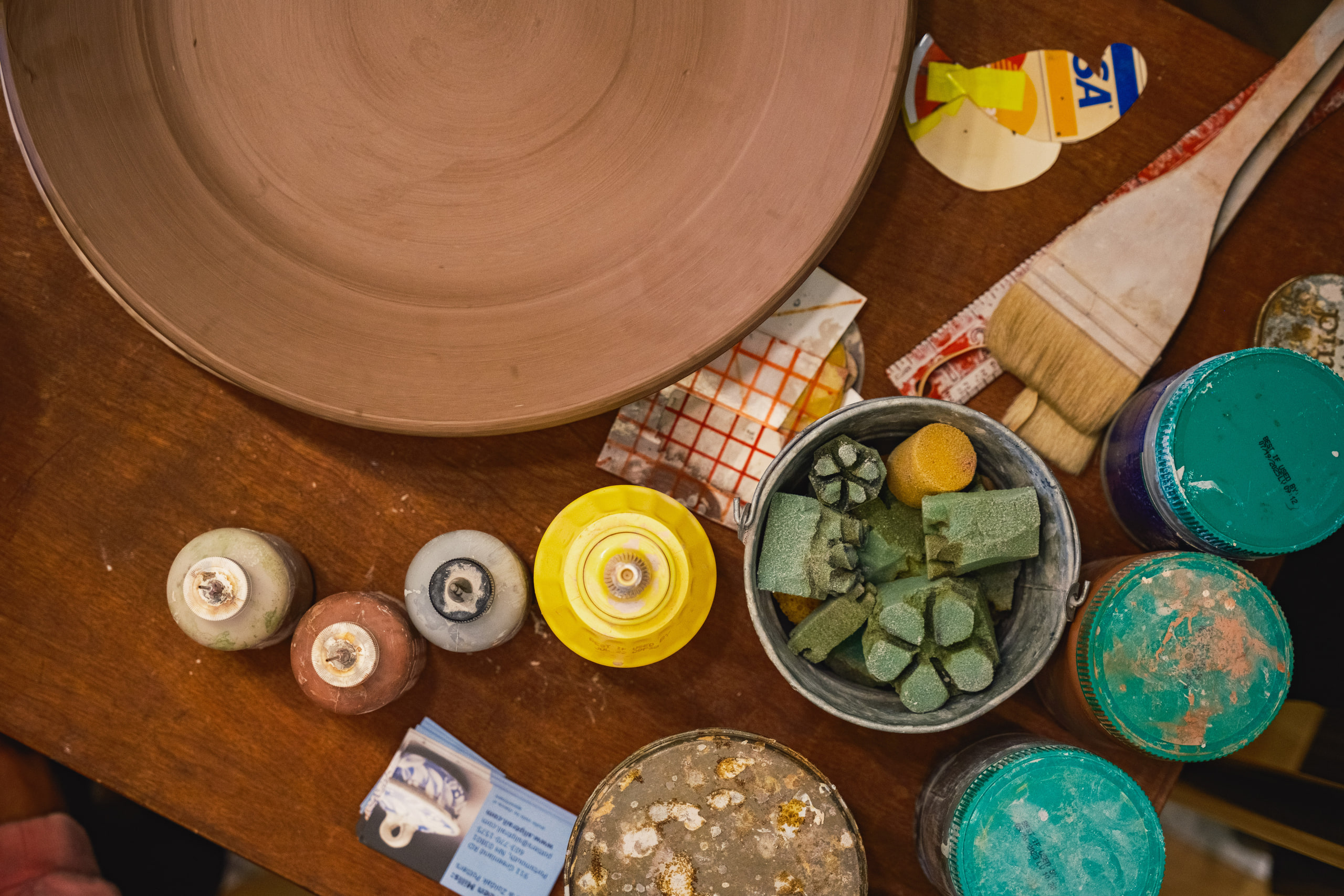 Looking down on a potter's worktable: an unfired clay plate, sponges in a tin, glaze pots and a wide brush.