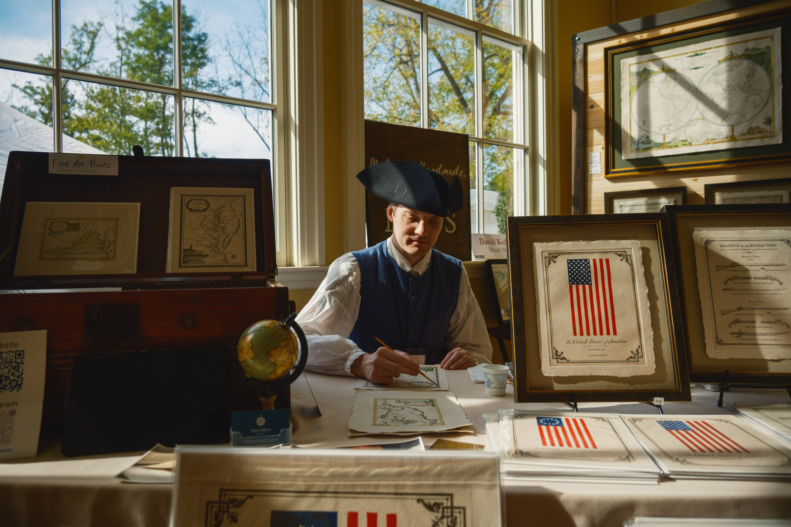 A man in a tricorn hat painting a map at a table by a sunny window, framed flag prints around him.