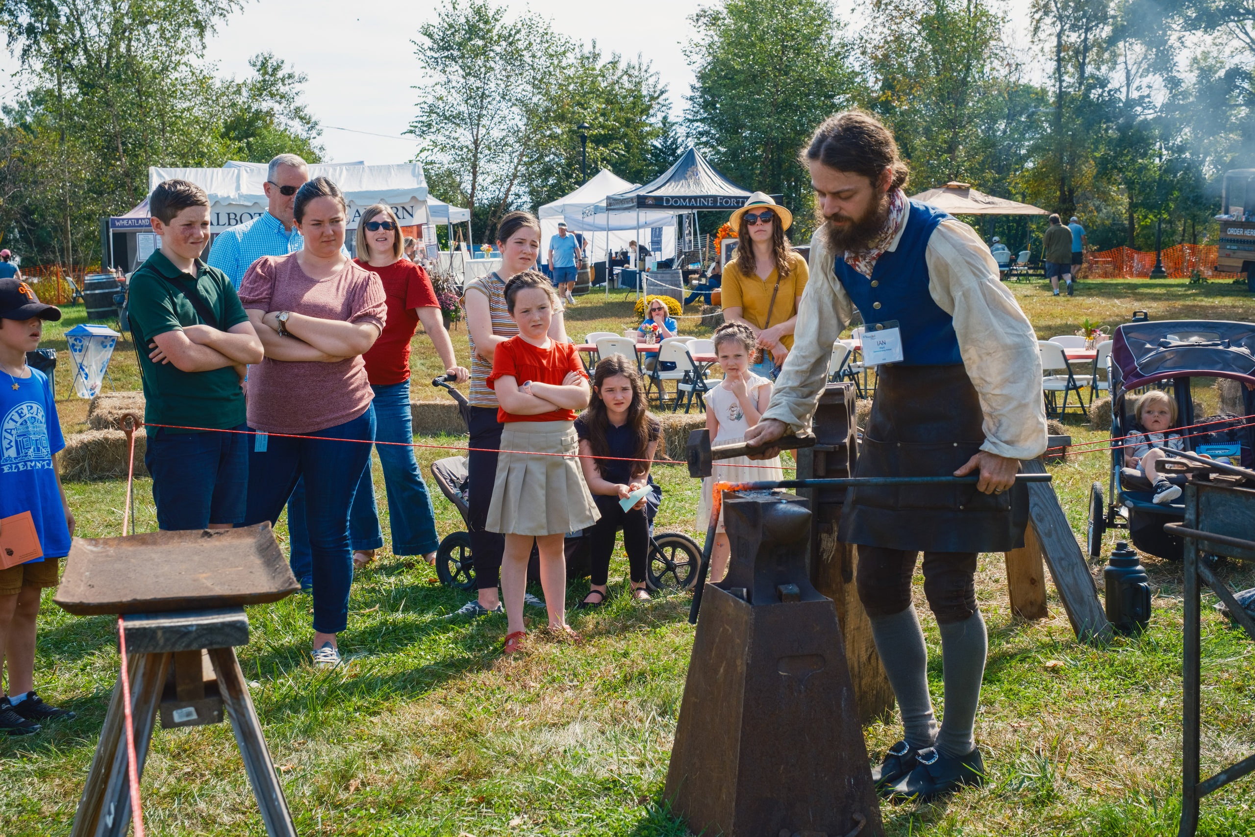 A blacksmith in period dress working iron at the anvil while a crowd watches from behind a rope at the Waterford Fair.