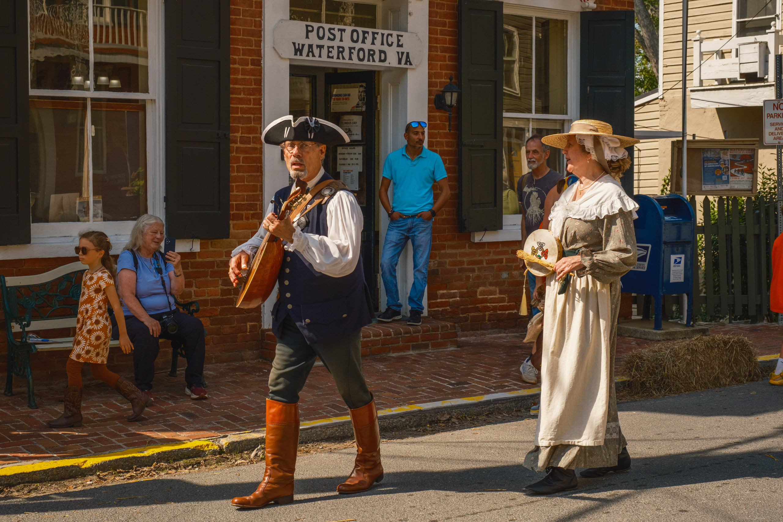 A musician in colonial dress playing a mandolin as he walks past the Waterford post office, a woman in period costume behind him, Waterford, Virginia.
