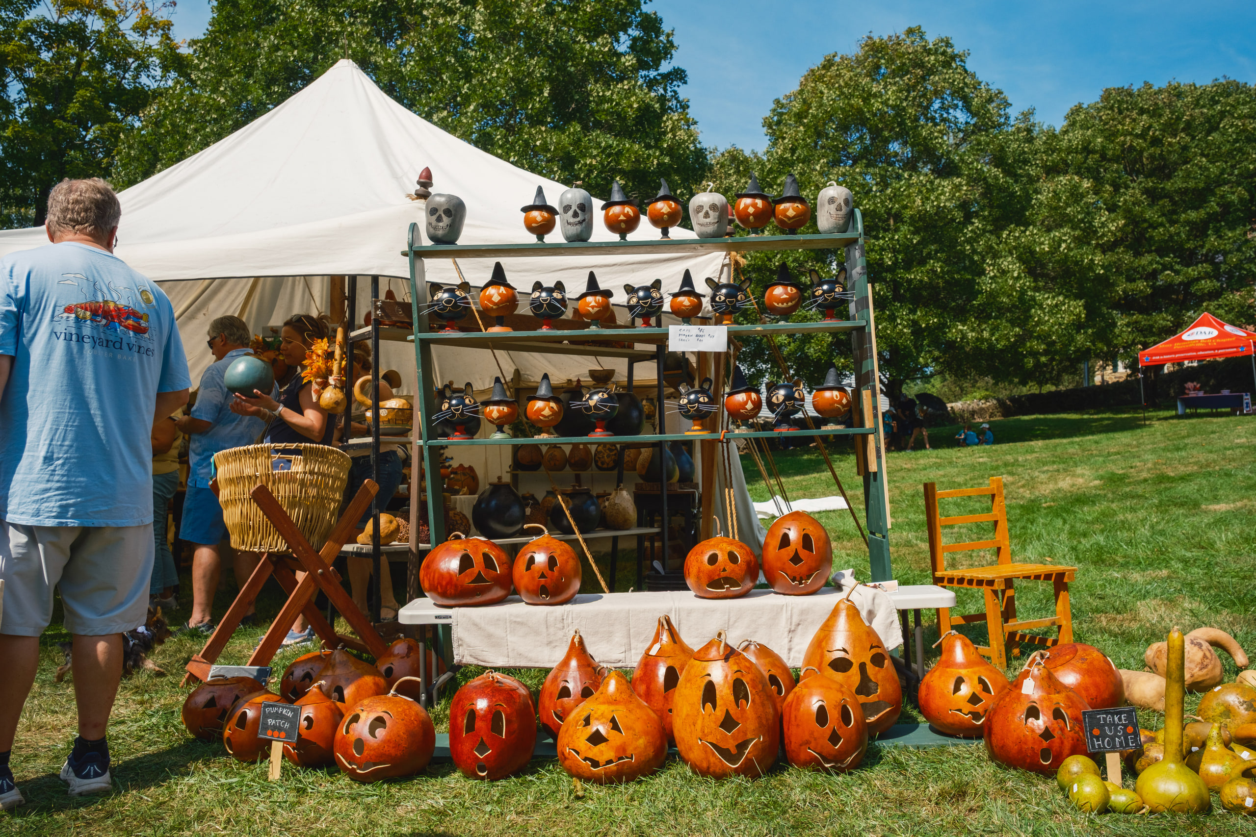 Carved gourd jack-o'-lanterns and painted gourds stacked on a craft booth on the grass in bright sun.