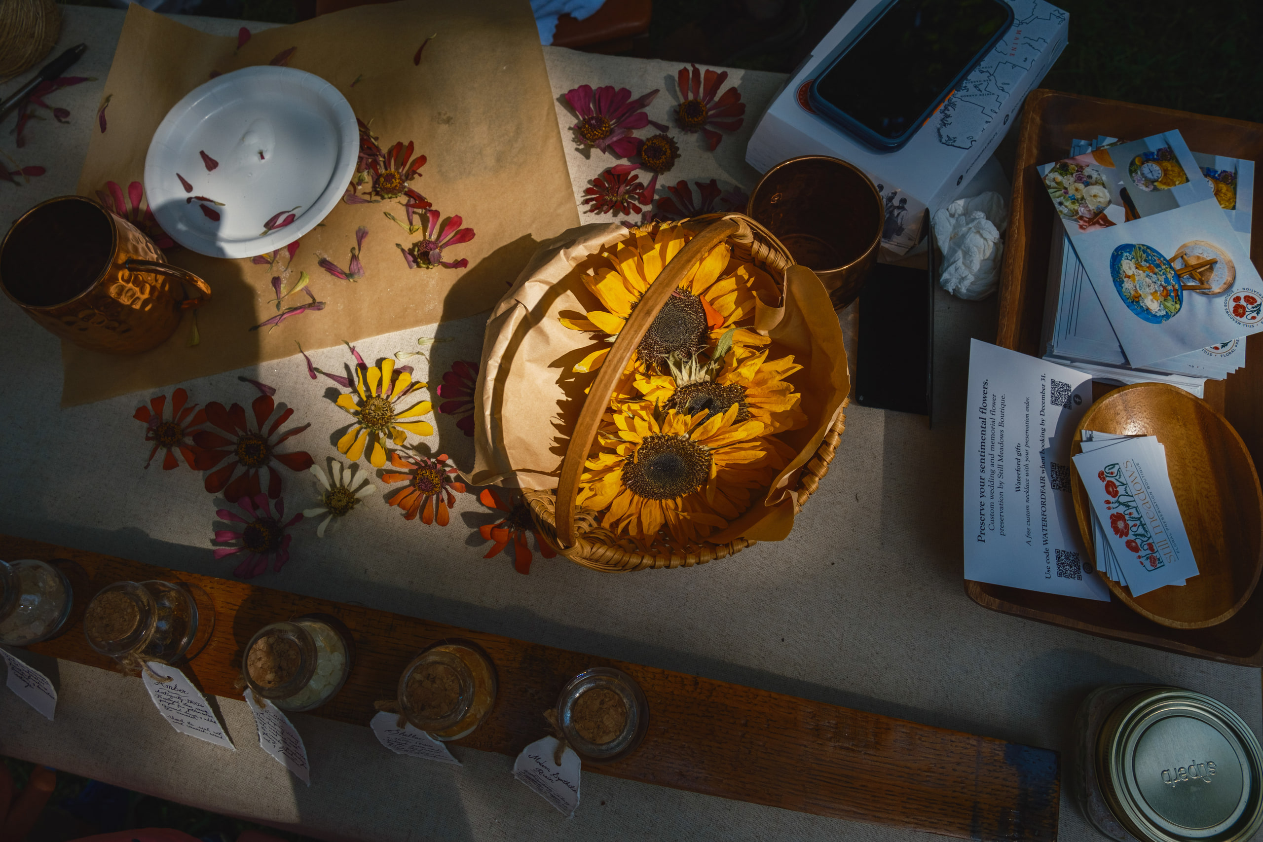 Looking down on a table of pressed flowers, a basket of sunflowers and small jars in late light.