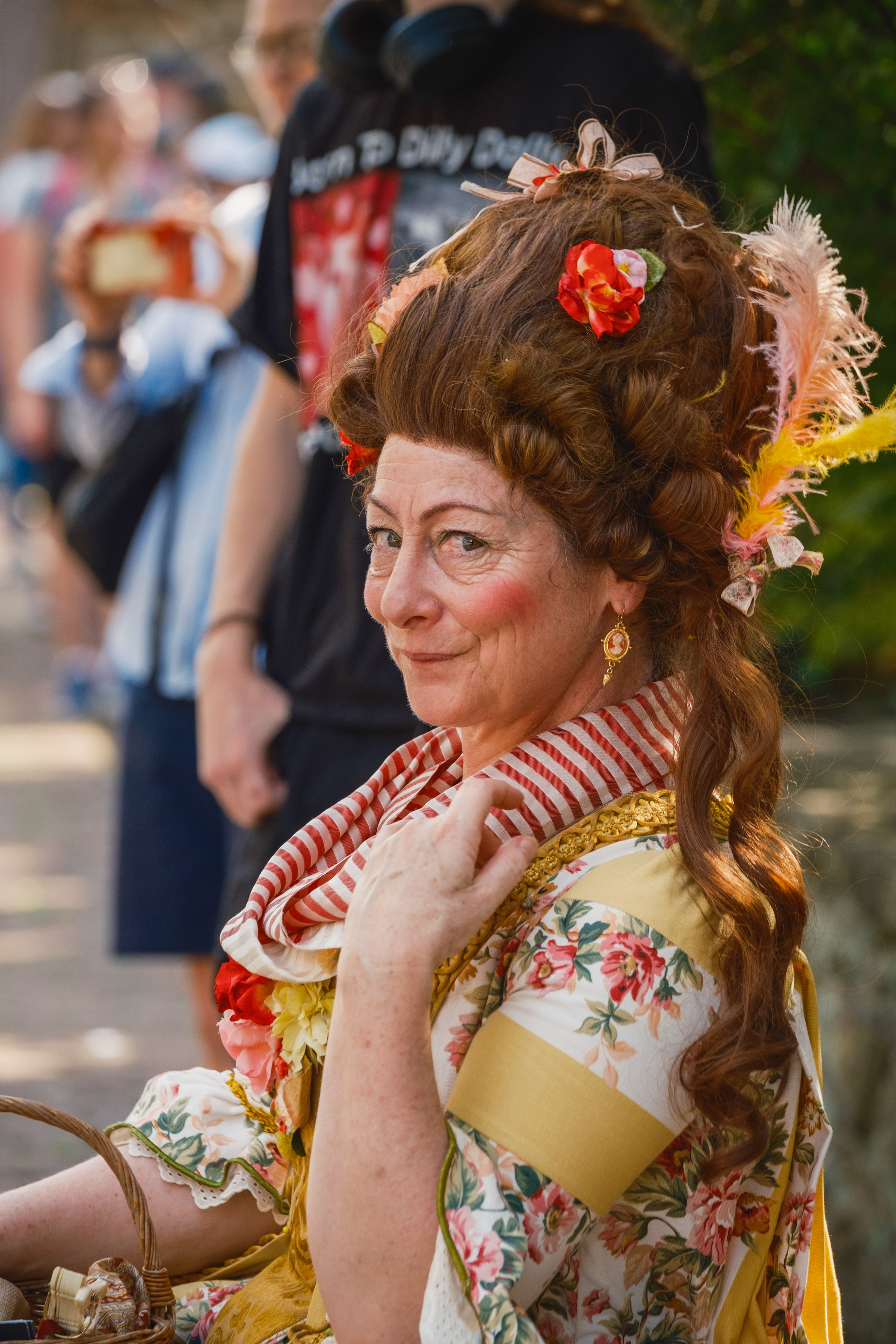 A woman in an 18th-century costume with curled hair, flowers and feathers, glancing back over her shoulder at the Waterford Fair.