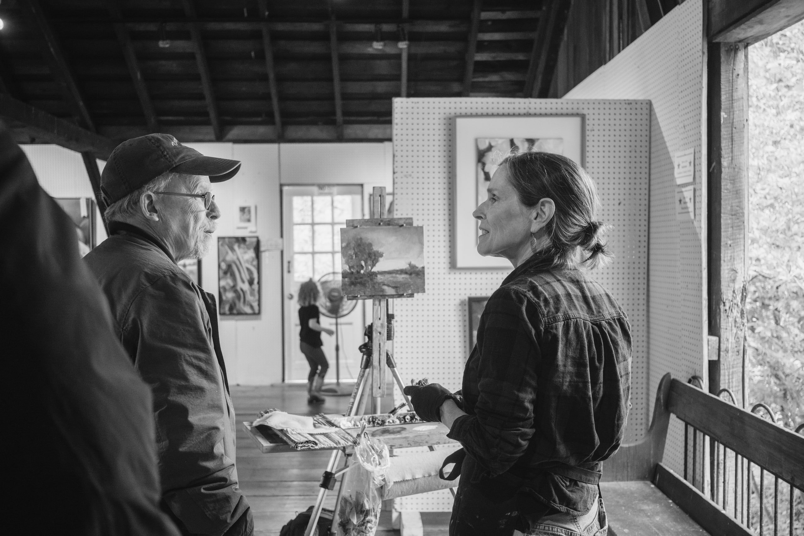 Black and white: a painter talking with a visitor in a barn gallery, her easel and a small landscape between them.