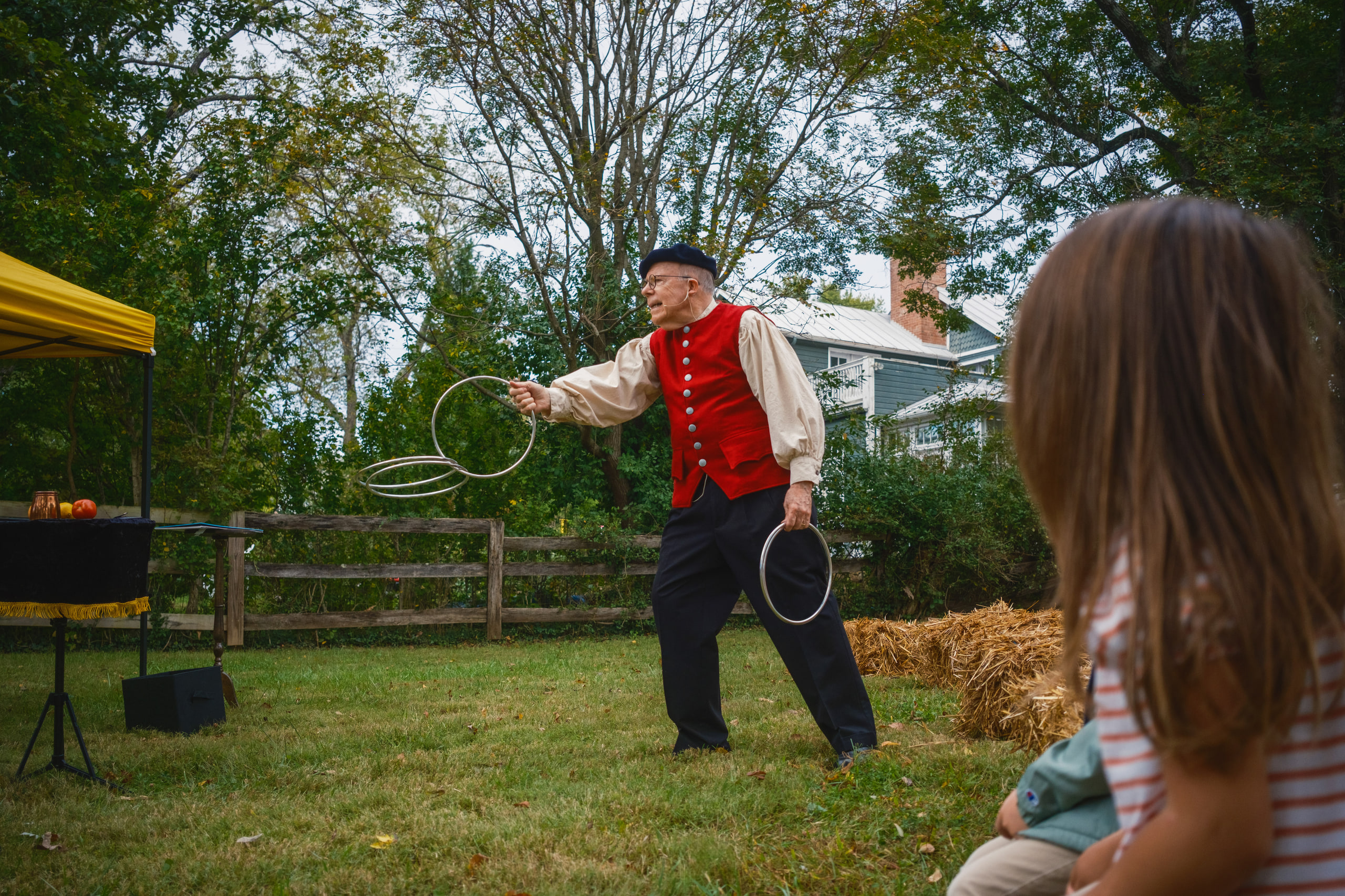 A performer in a red waistcoat throwing linking metal rings on a lawn, the audience watching from behind.