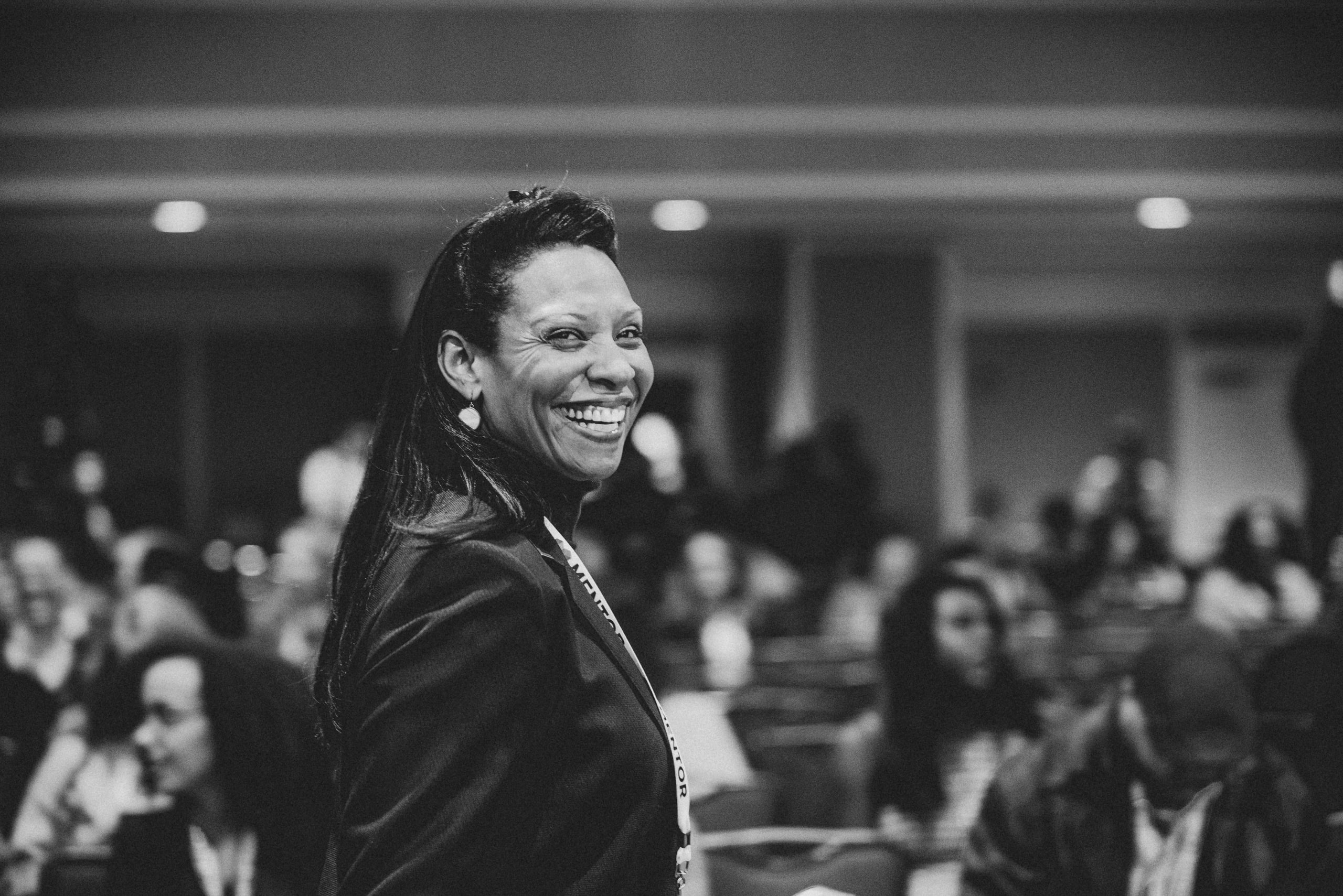 Black and white: a woman with a lanyard laughing in a crowded conference room.