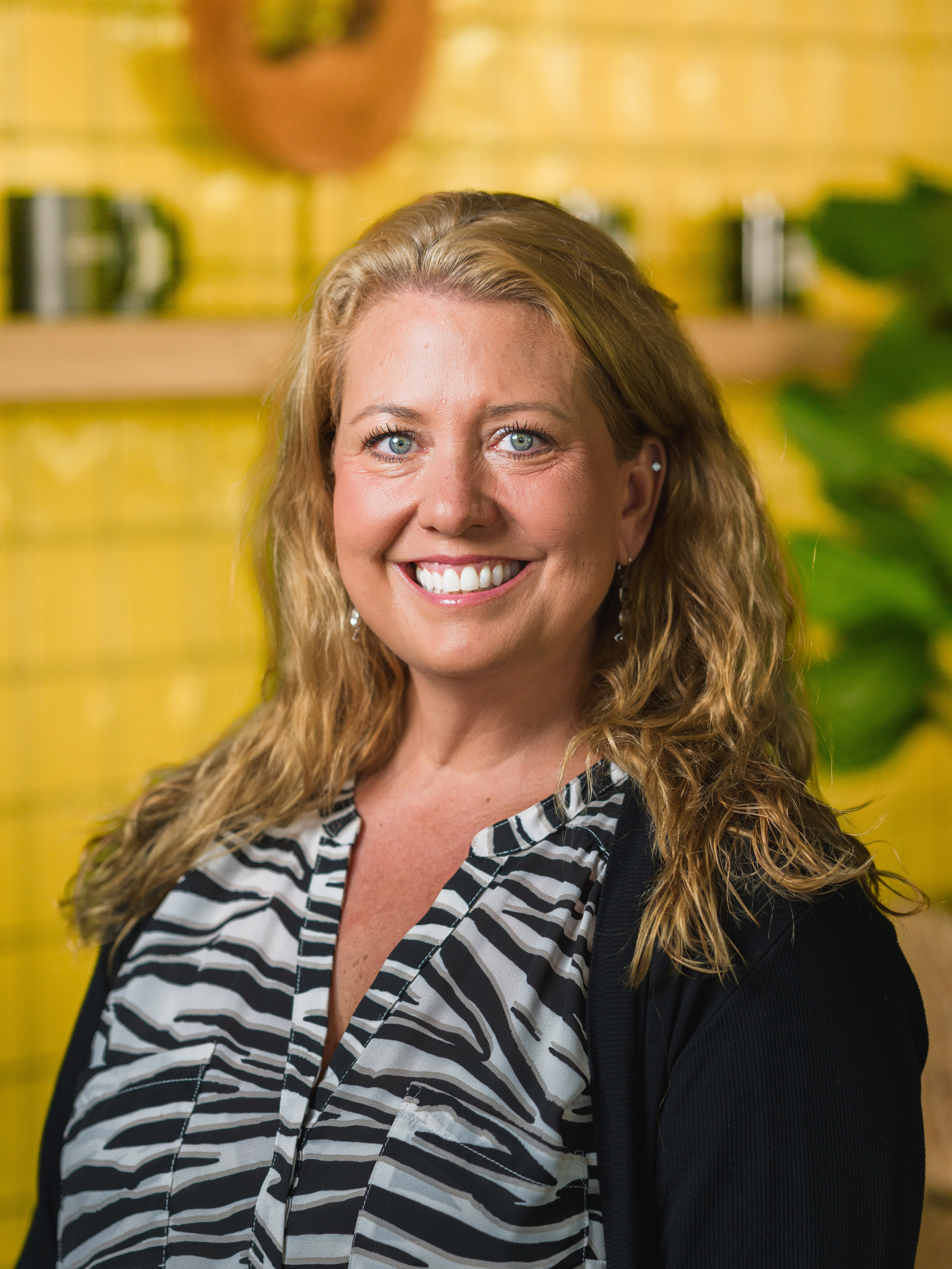 Portrait of a smiling woman with long blonde hair in a zebra-print blouse against a yellow tile wall, Ashburn, Virginia.