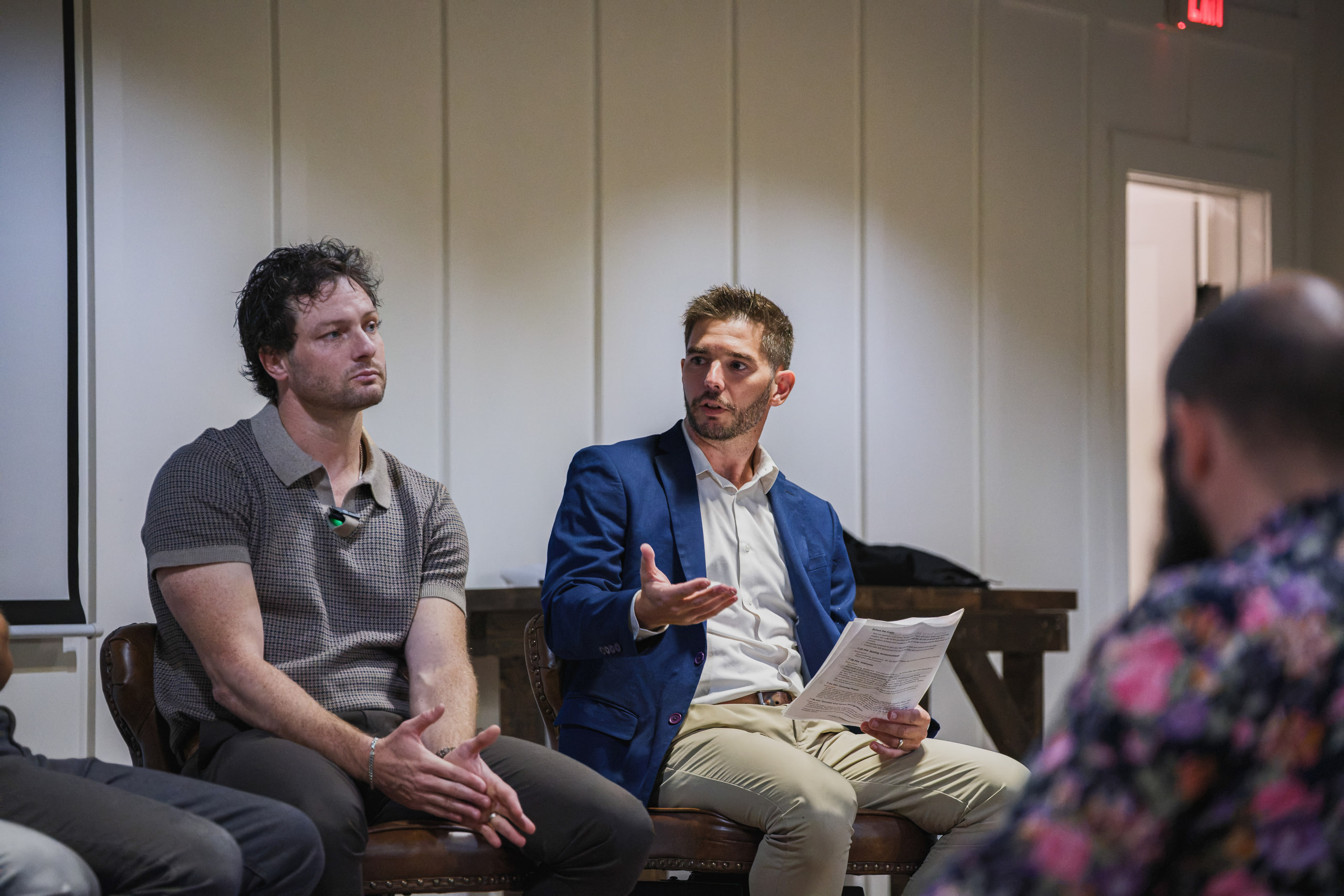 Two panelists on stools, one in a blue blazer answering with notes on his knee.