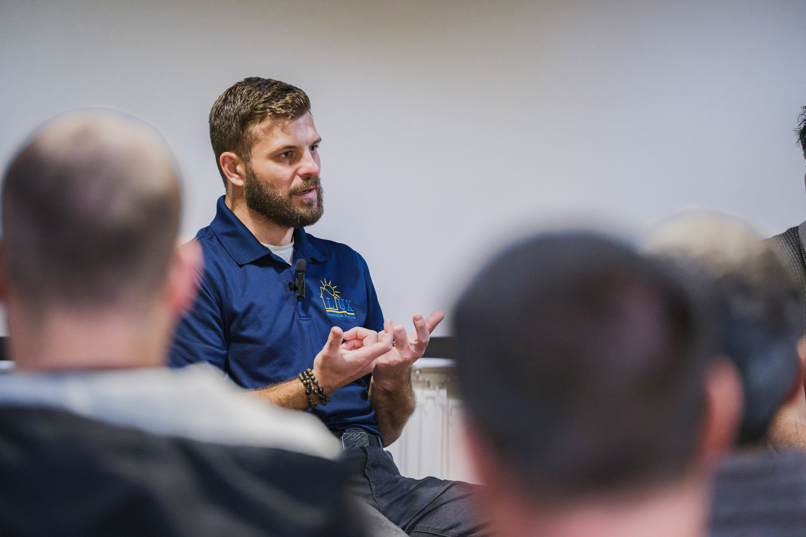 A bearded panelist in a blue polo gestures as he speaks, seen past the heads of the audience.
