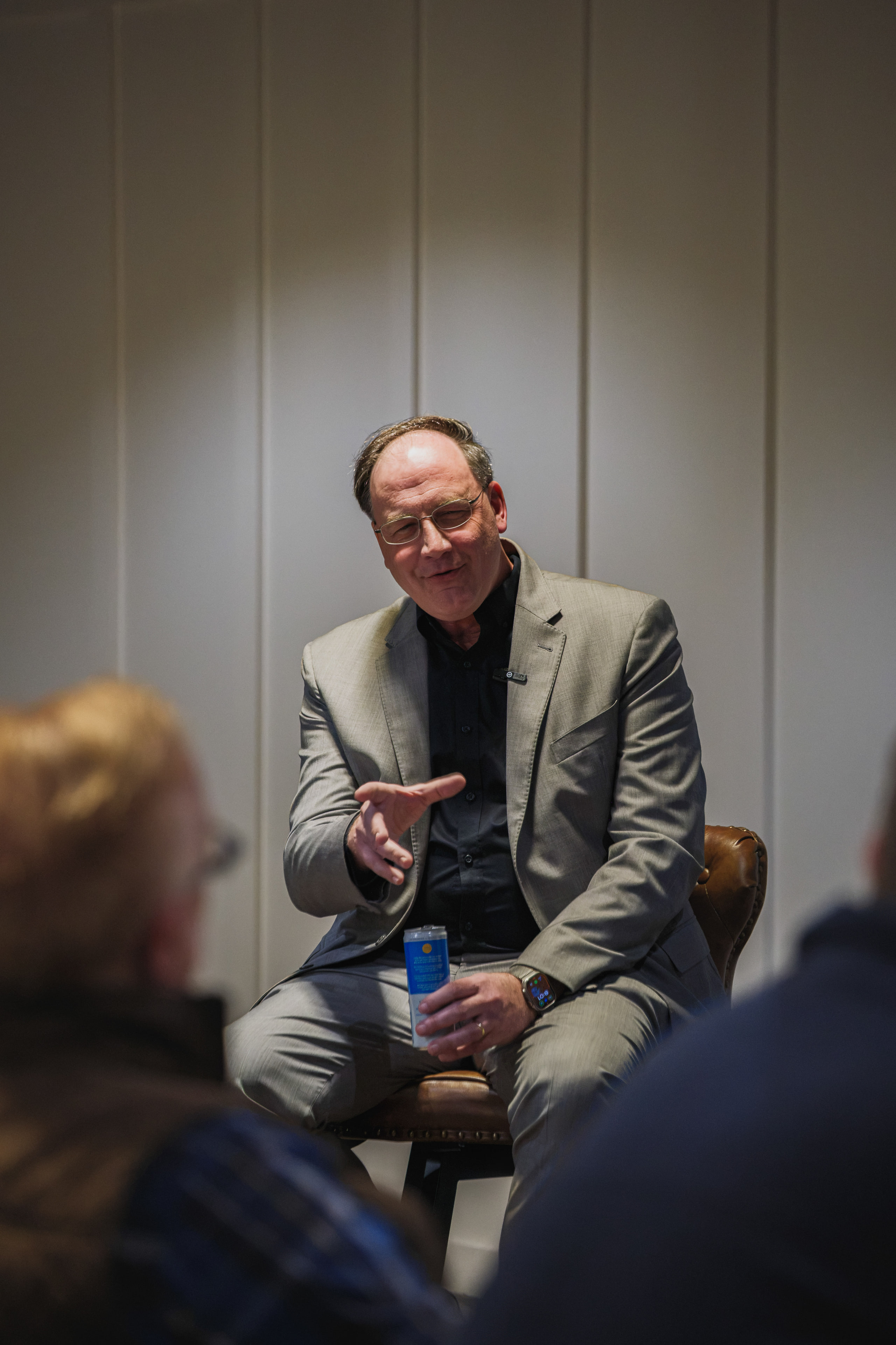 A speaker in a gray blazer leans forward mid-answer on a leather chair, a can in one hand.