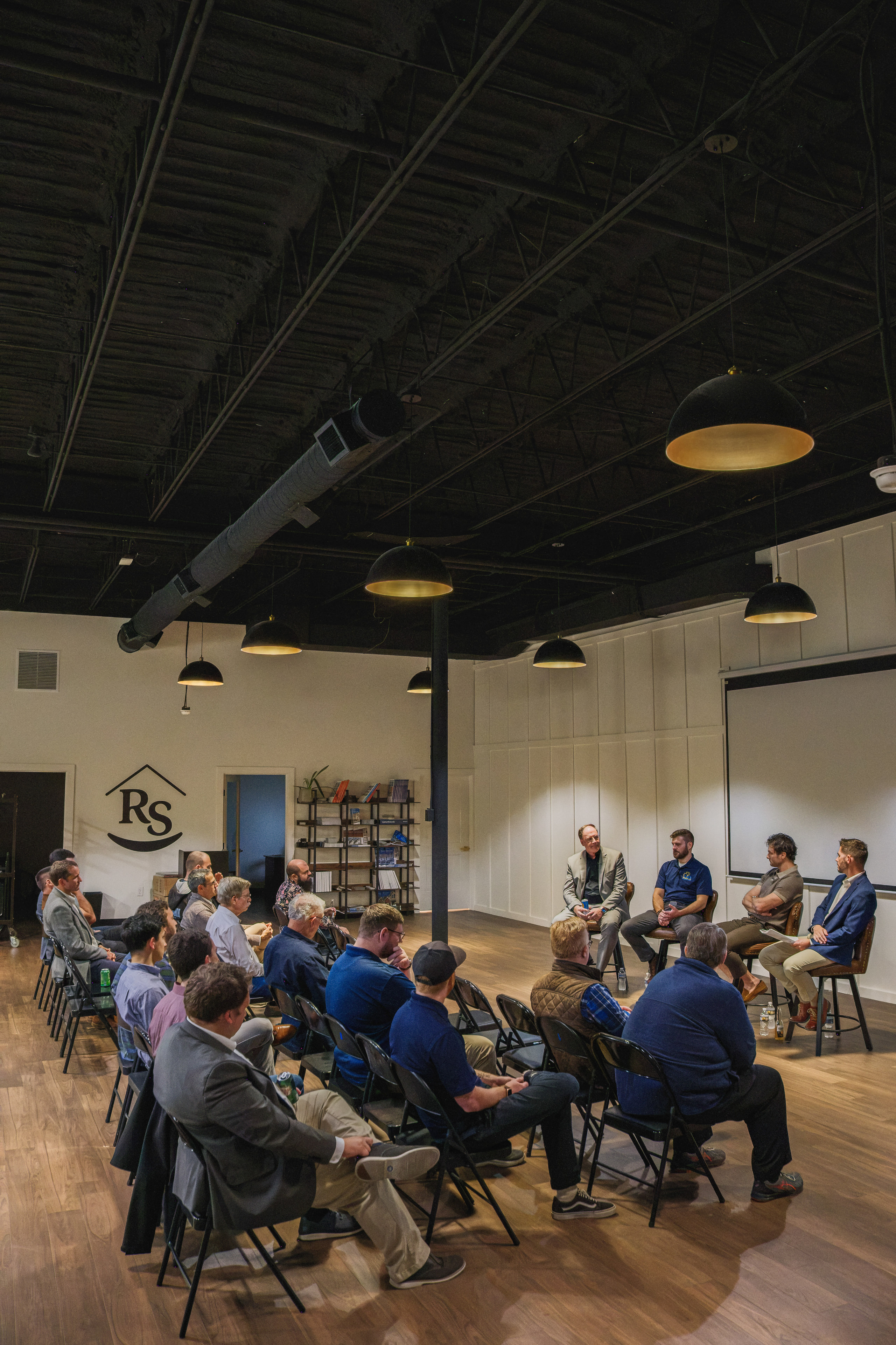 An evening panel seen from above: a full circle of chairs facing three speakers under warm pendant lights.