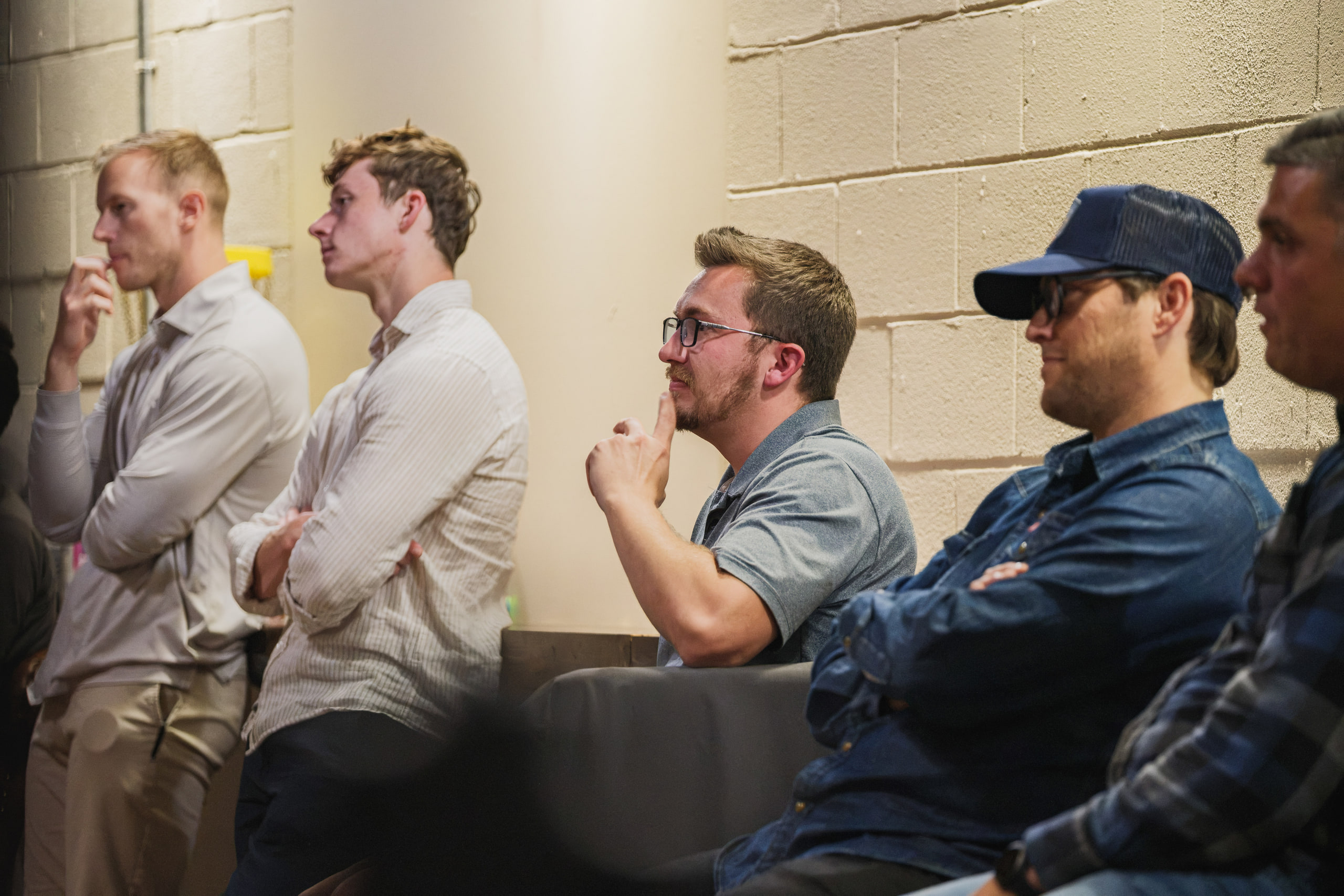 Audience members listen along a block wall, one with a hand at his chin.