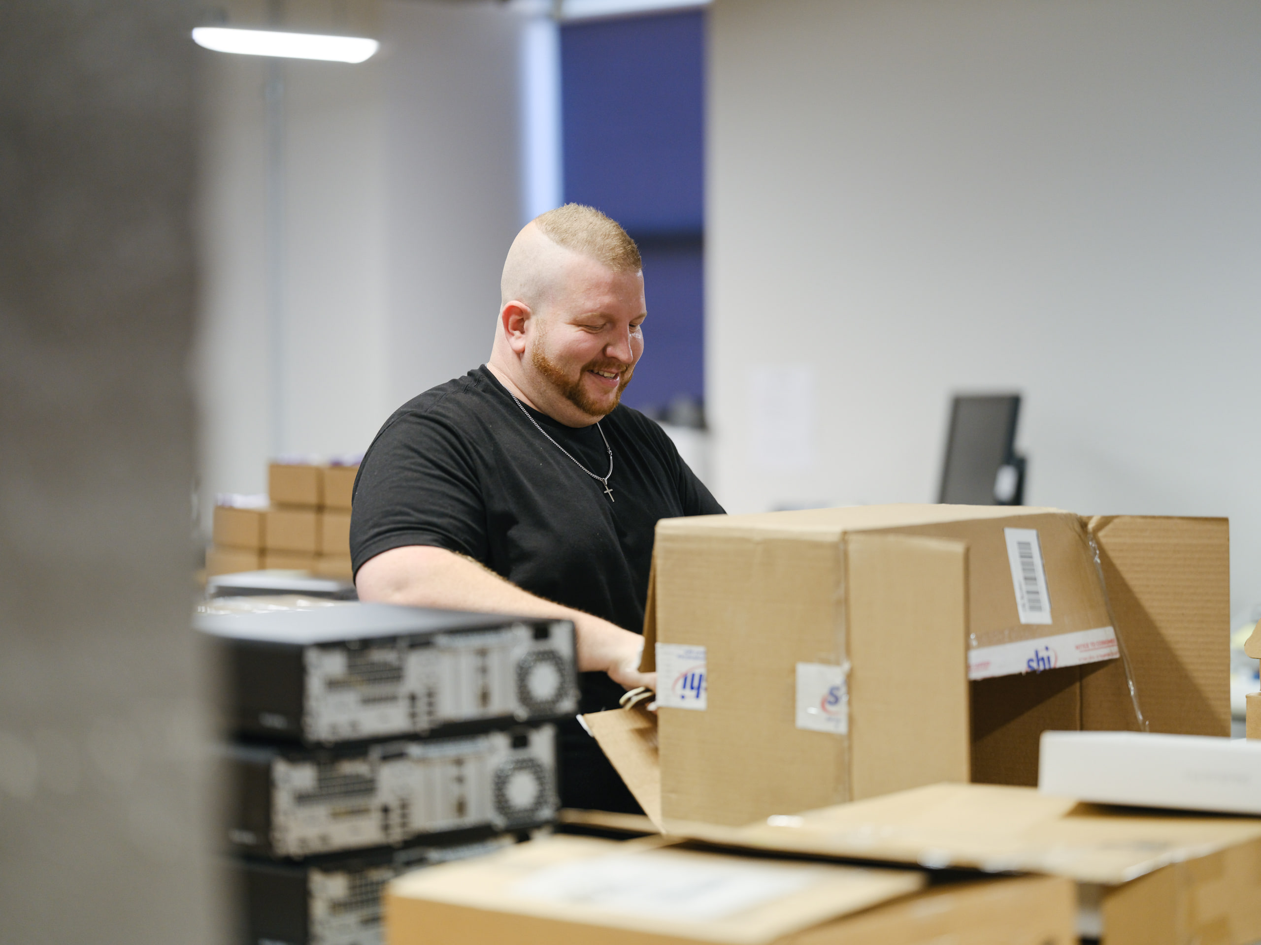 A smiling man in a black t-shirt opens a cardboard shipping box in a bright warehouse, with stacked computer towers in the foreground.