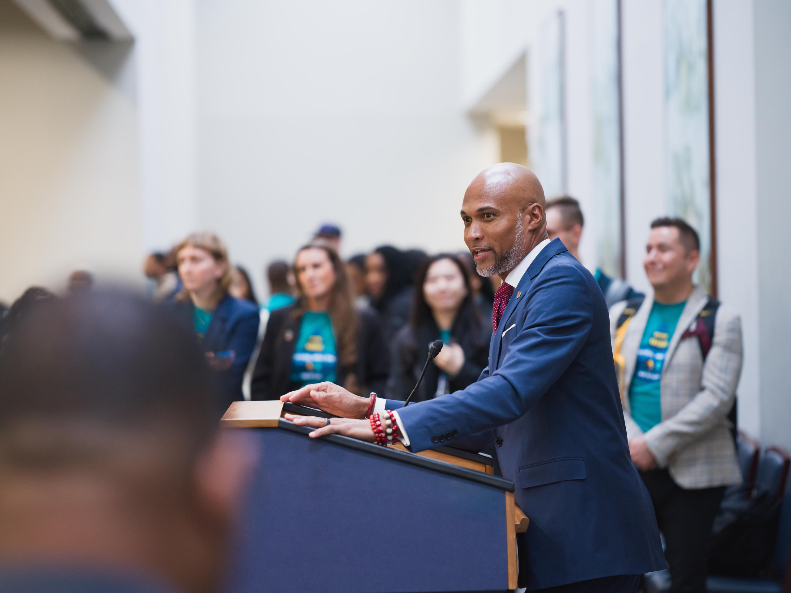 Speaker leaning into the crowd, teal shirts behind at the Capitol Hill reception in Washington, DC.