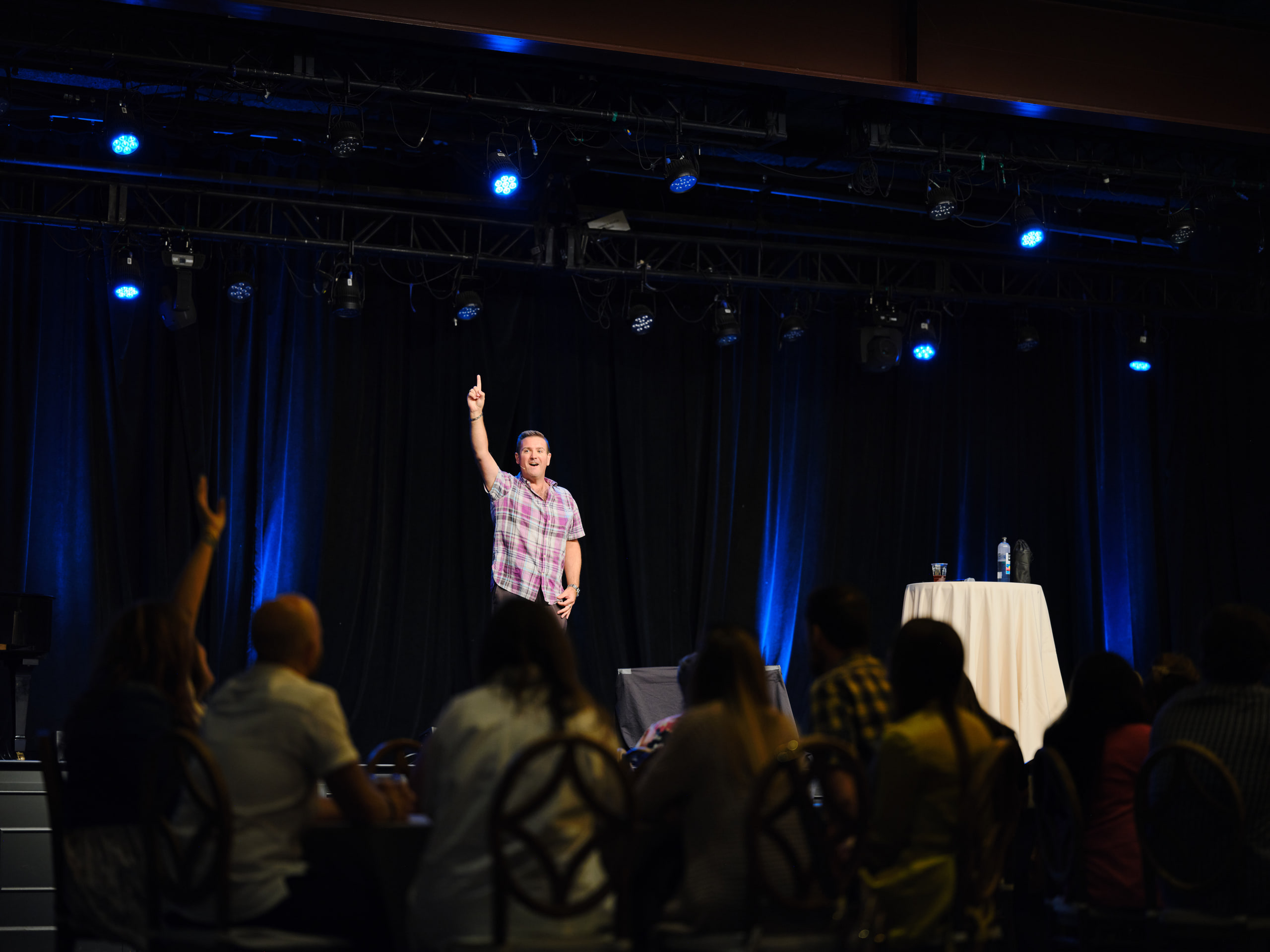 Speaker on stage, arm up, blue at a team-building event in a brick warehouse venue, Nashville.