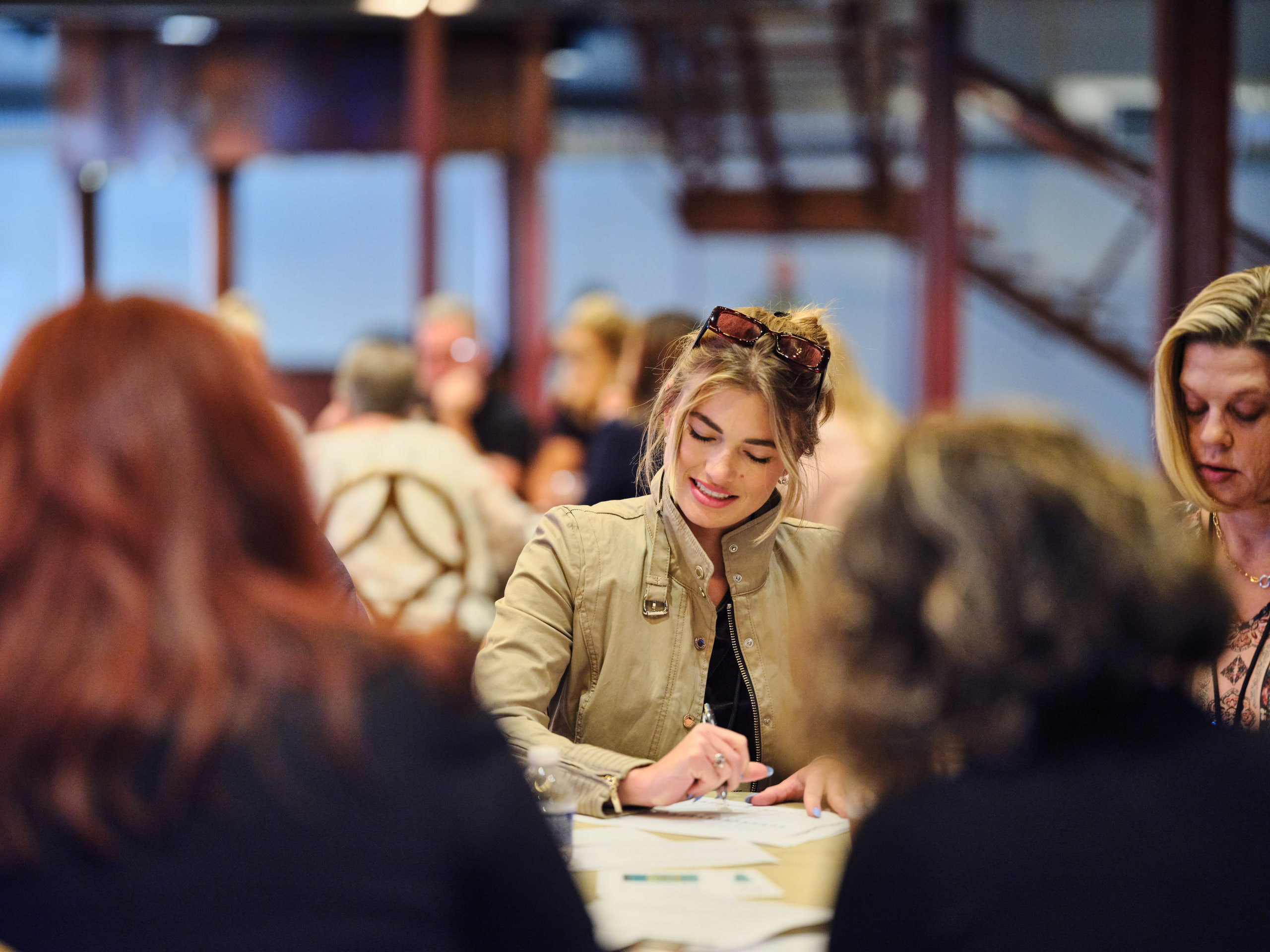 A woman smiling as she writes at a table during a team session, colleagues blurred around her in a warm hall