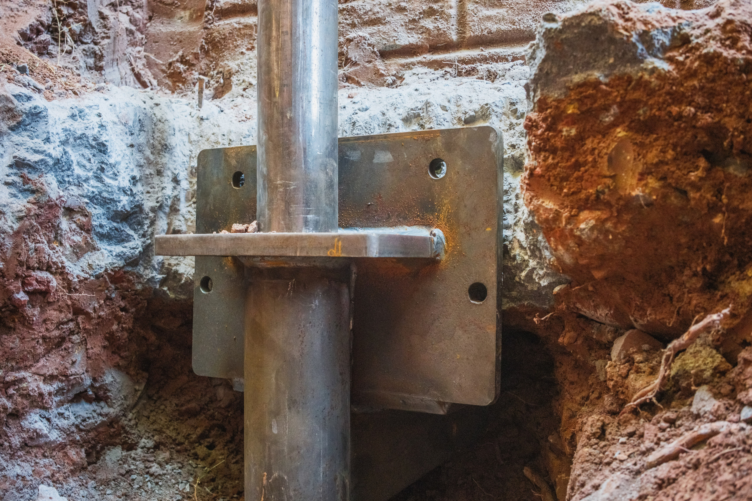 A steel pier and bracket set under a footing in red clay.