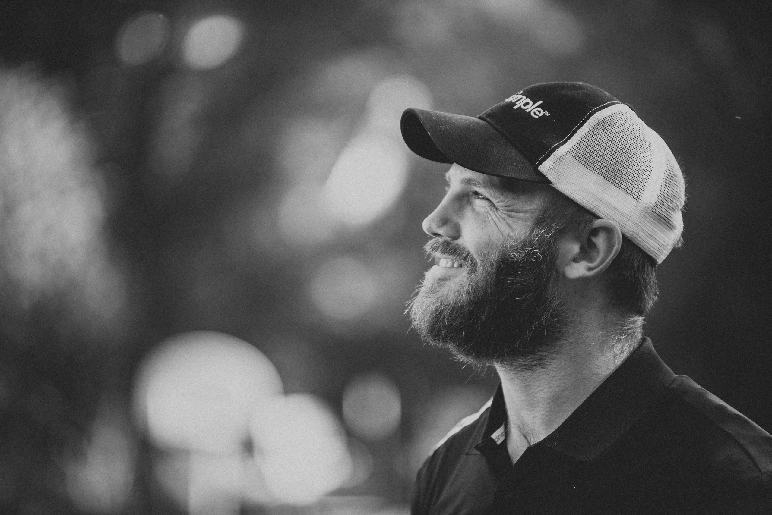Black and white profile of a bearded man in a trucker cap smiling and looking up, with soft circles of out-of-focus light filling the left of the frame.