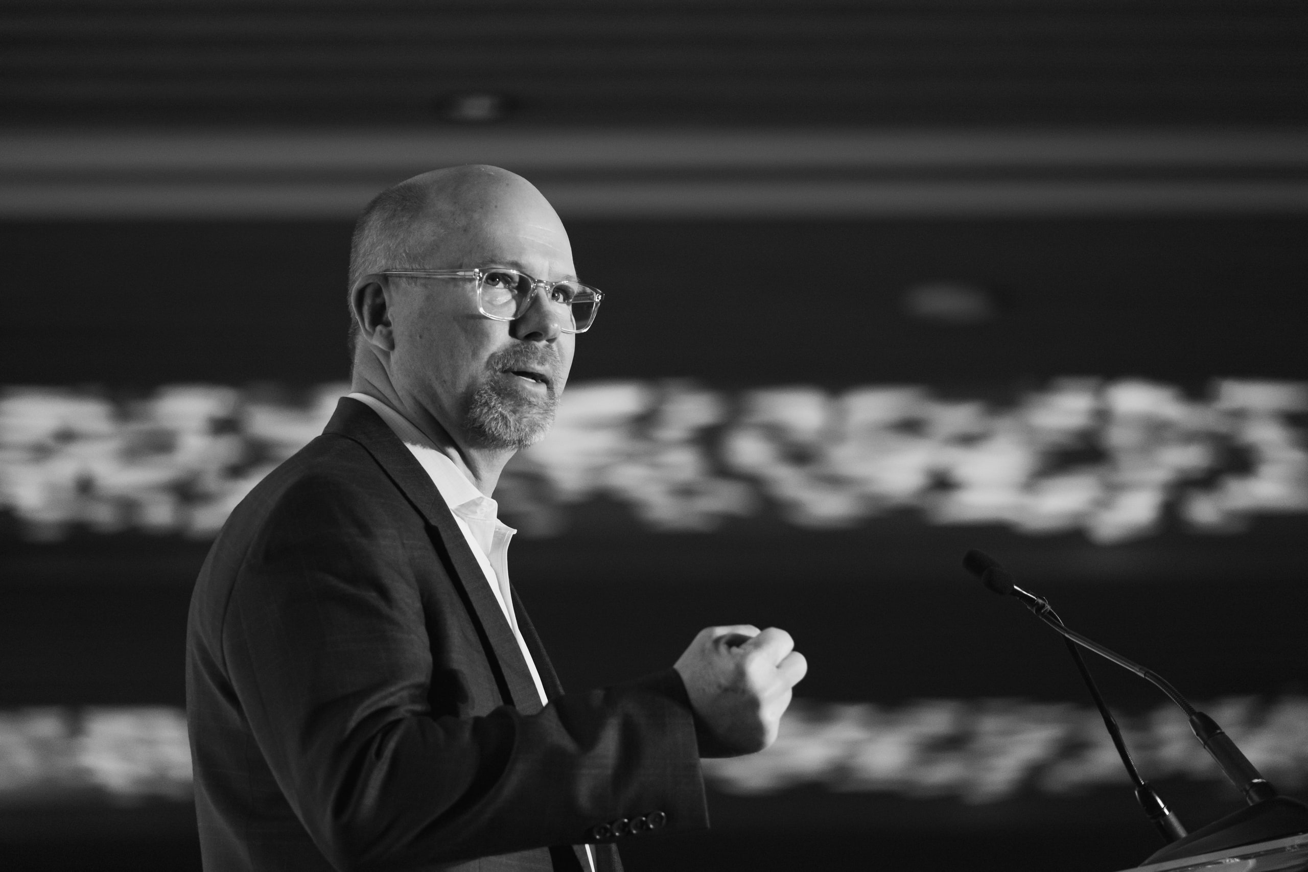 Black and white speaker with glasses, ceiling pattern at a national summit plenary.