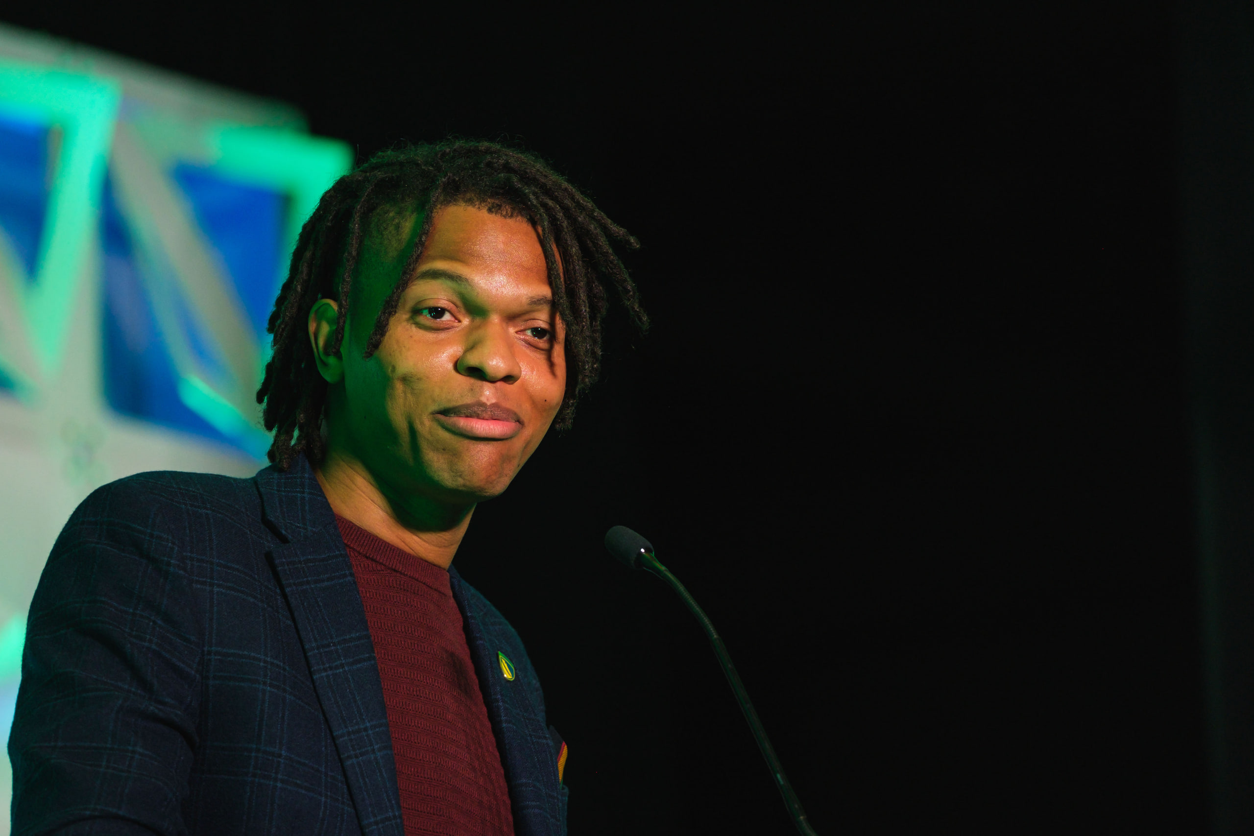 A young speaker smiling at the microphone against a dark stage.