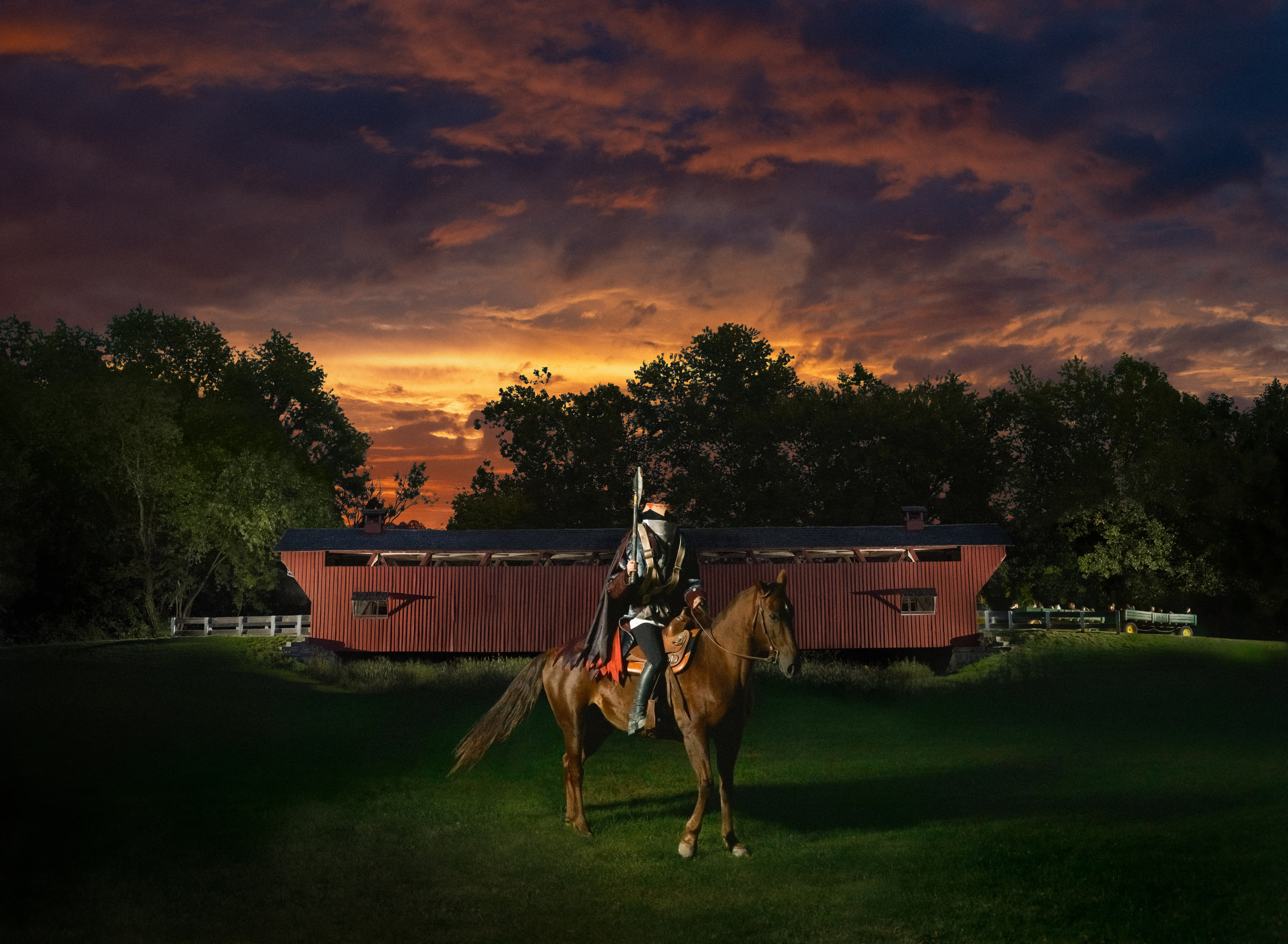 A rider in armour on a chestnut horse in front of a red covered bridge under a fiery sunset sky.