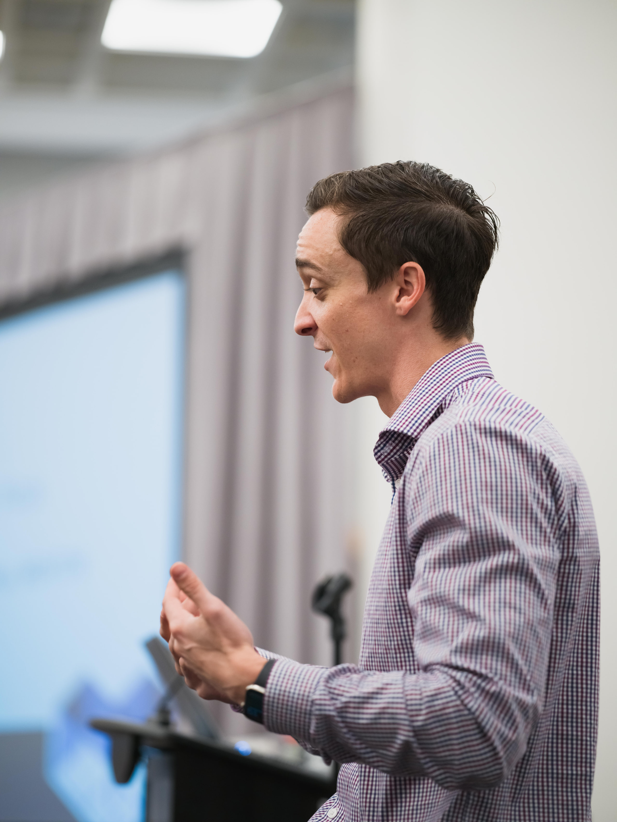 A speaker in a lavender shirt talking with his hands beside a lectern and a screen.