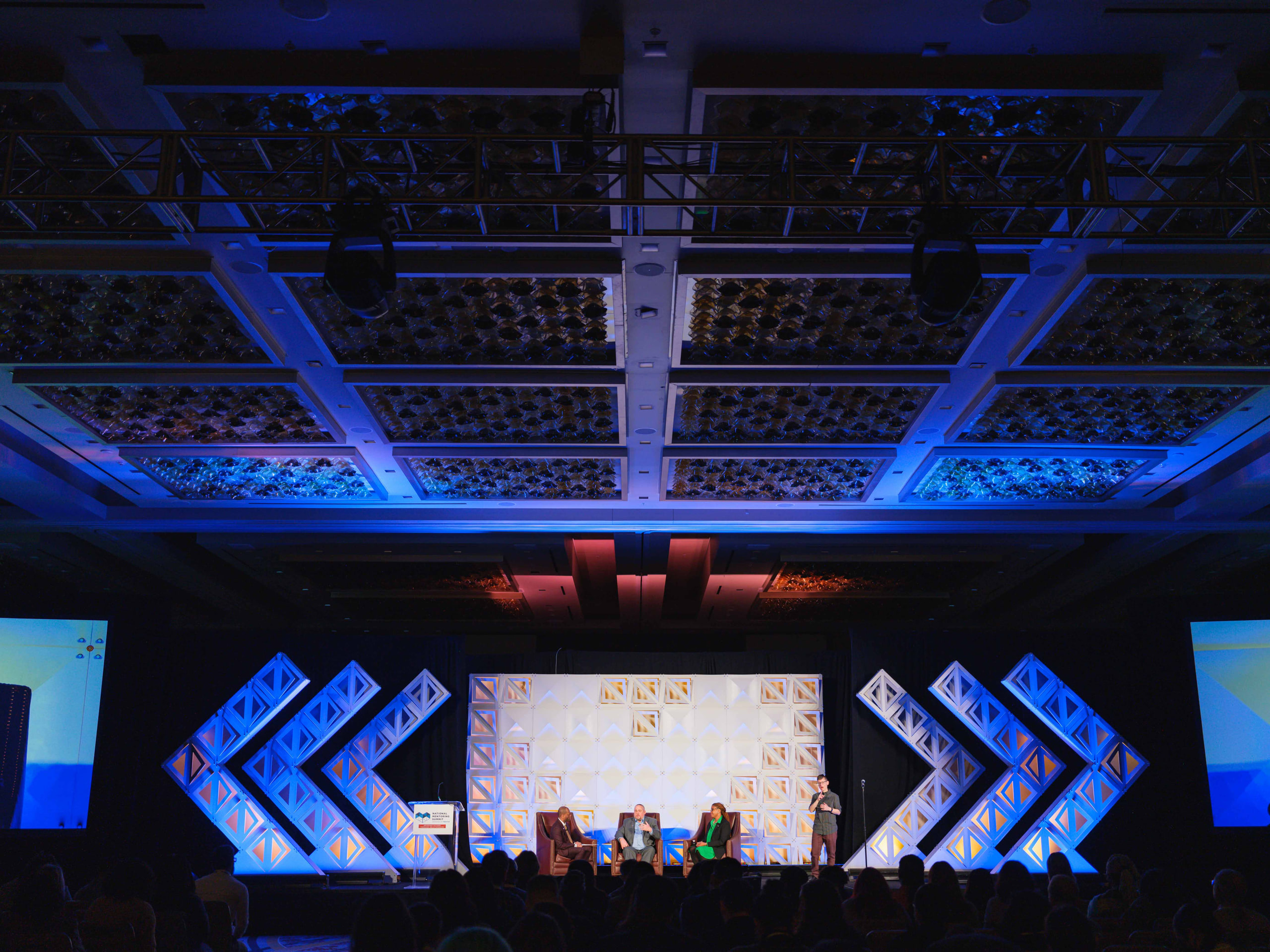 A wide view of a ballroom with blue chevron screens around a stage and a full audience.
